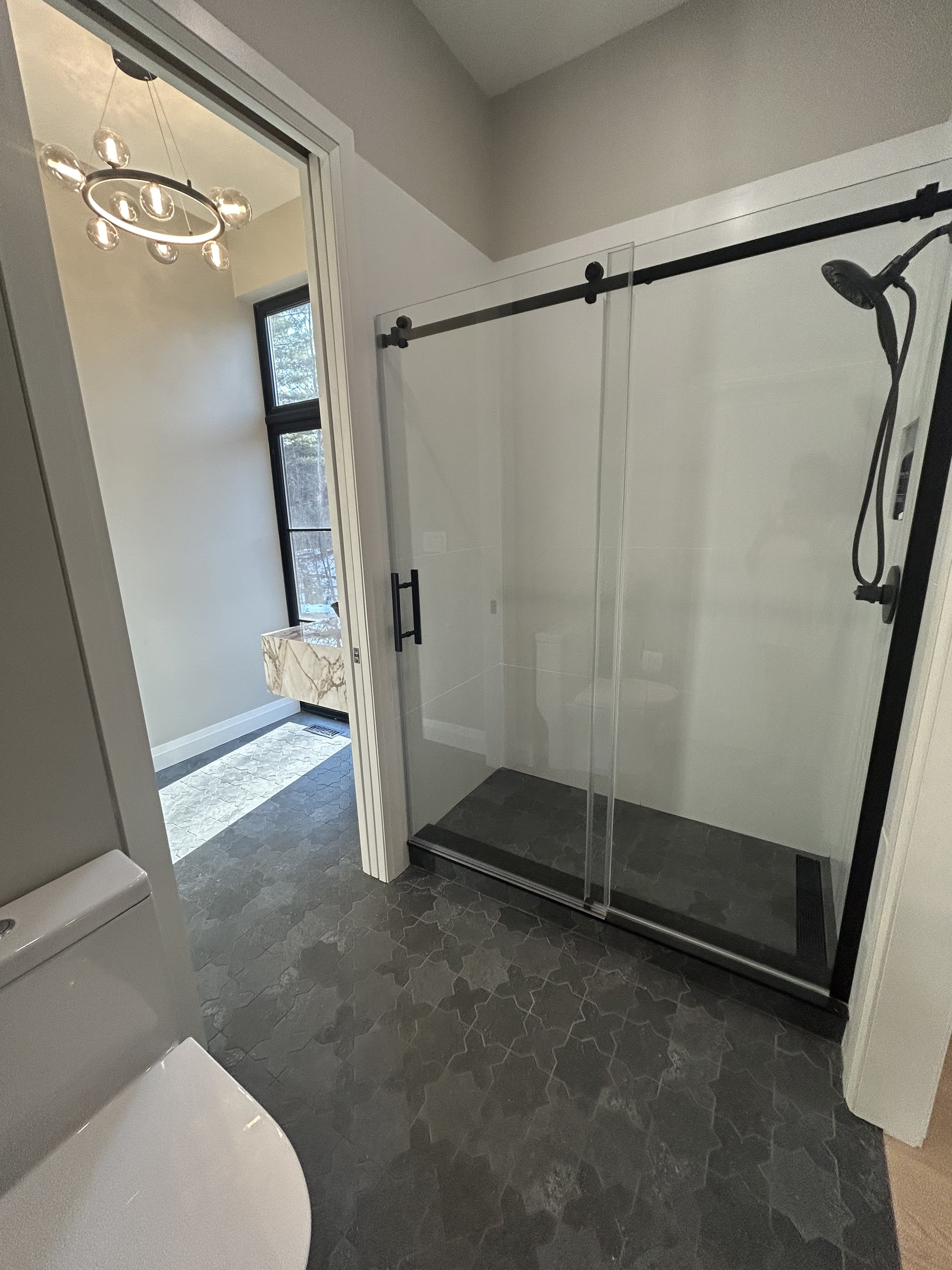 Bathroom renovation with a glass shower enclosure, a toilet, and a window with a view of trees outside. There is a marble sink visible through the doorway and a modern ceiling light fixture.