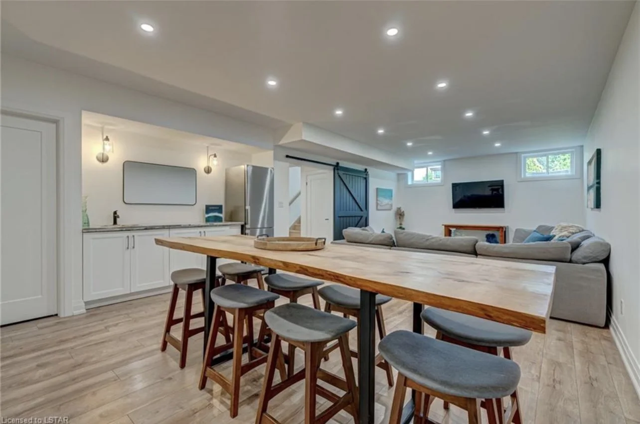 Open-concept basement with a kitchen area on the left featuring white cabinets, a stainless steel refrigerator, and a large mirror. seating area with a large gray sectional sofa, TV on the wall, and egress window near the ceiling for natural light.