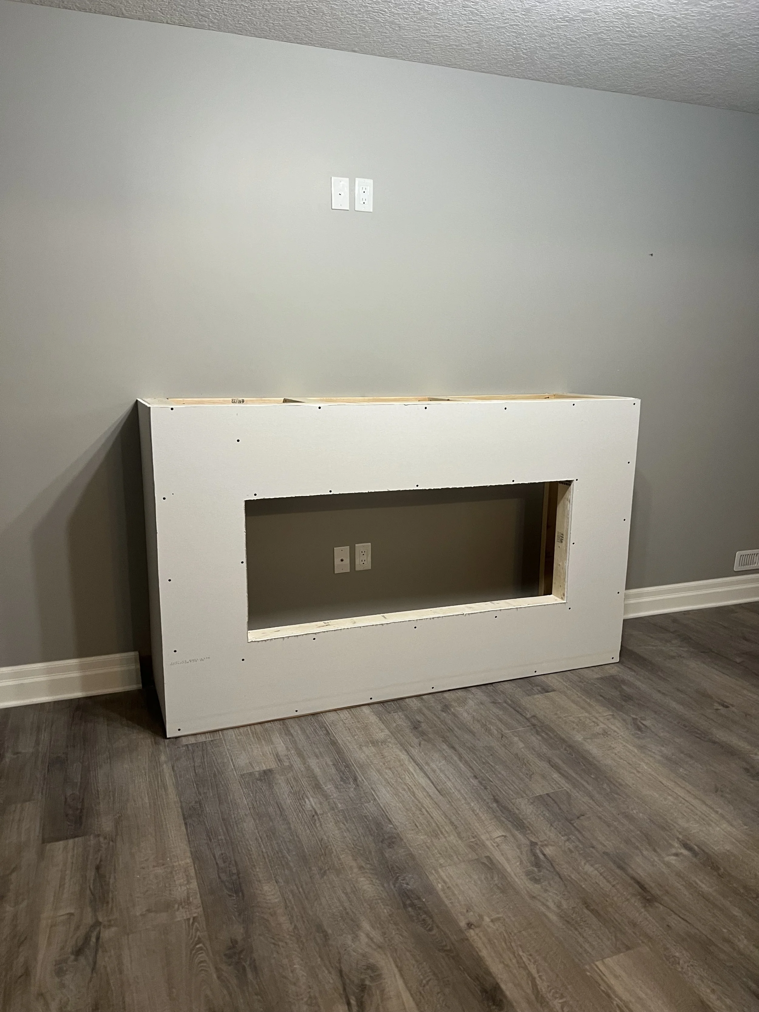 Unfinished white wooden frame against a gray wall on a hardwood floor, with electric outlets visible inside the frame in a basement renovation.
