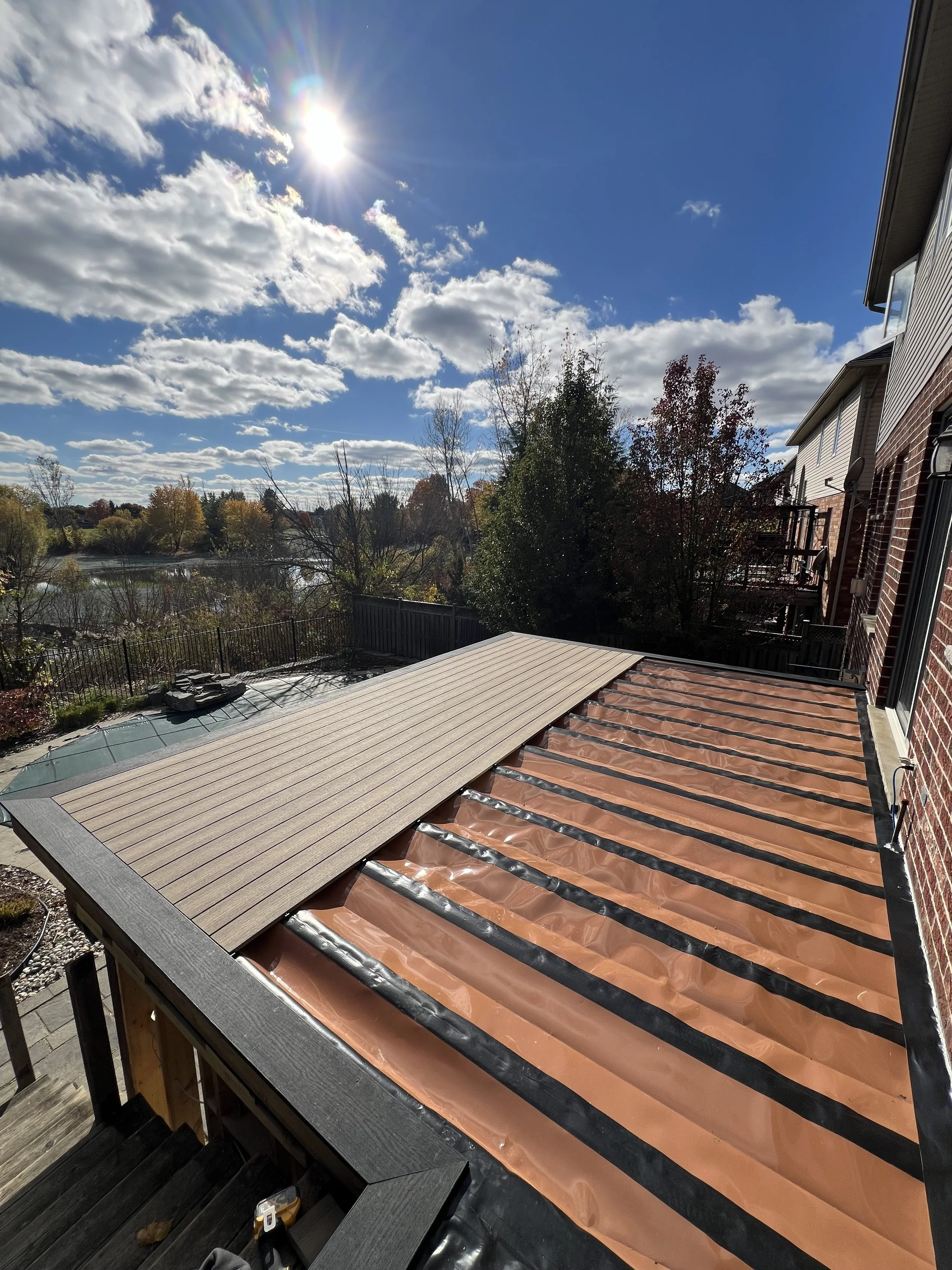 The image shows a partial view of a composite deck with  new beige-colored decking and exposed water proofing In the background, there are trees with autumn foliage, a body of water, and a partly cloudy blue sky with the sun shining brightly.