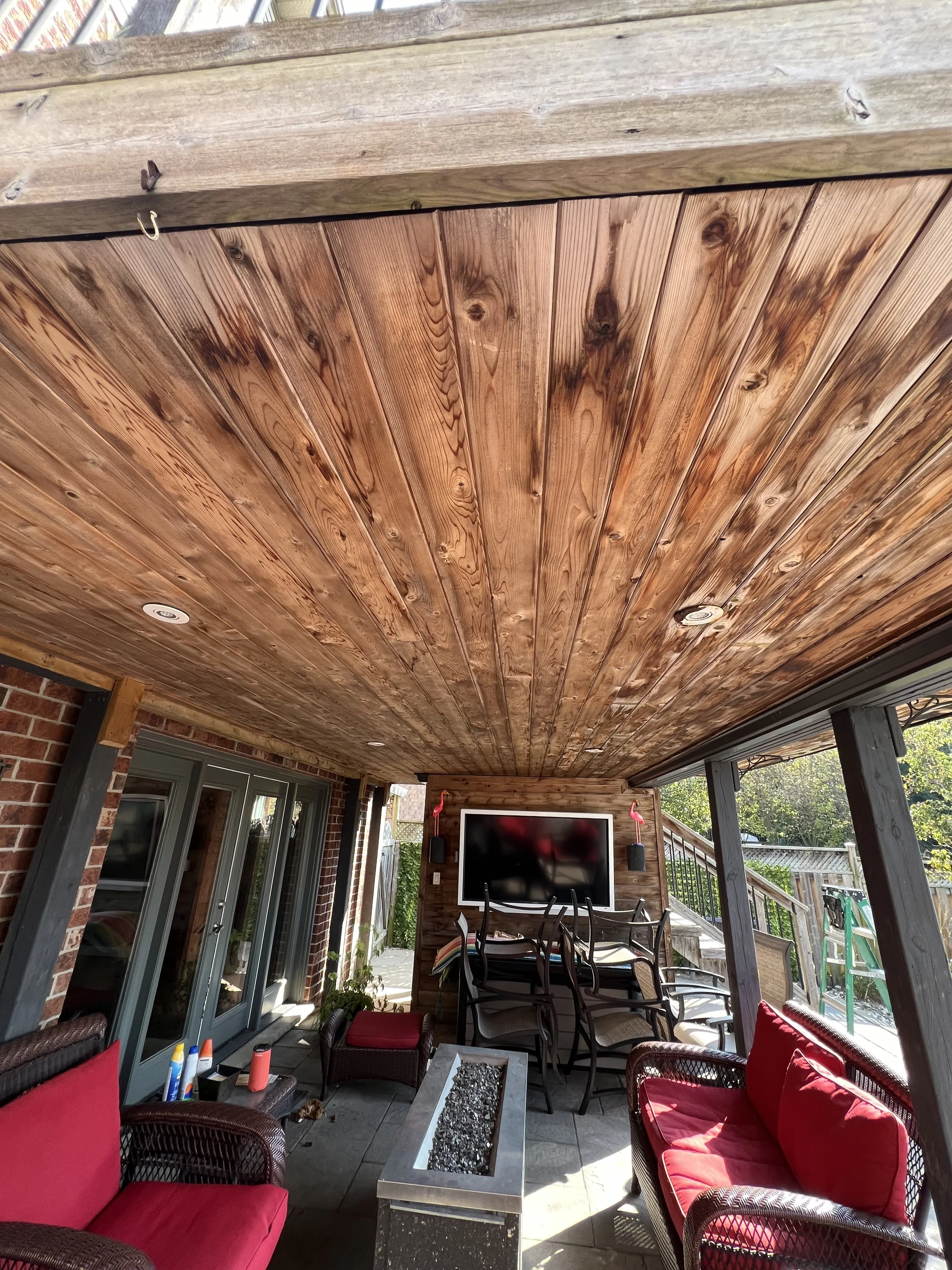 Covered outdoor patio with red cushioned wicker furniture, a rectangular fire pit table, a mounted flat-screen TV, stacked chairs, and string lights, adjacent to a brick house wall.