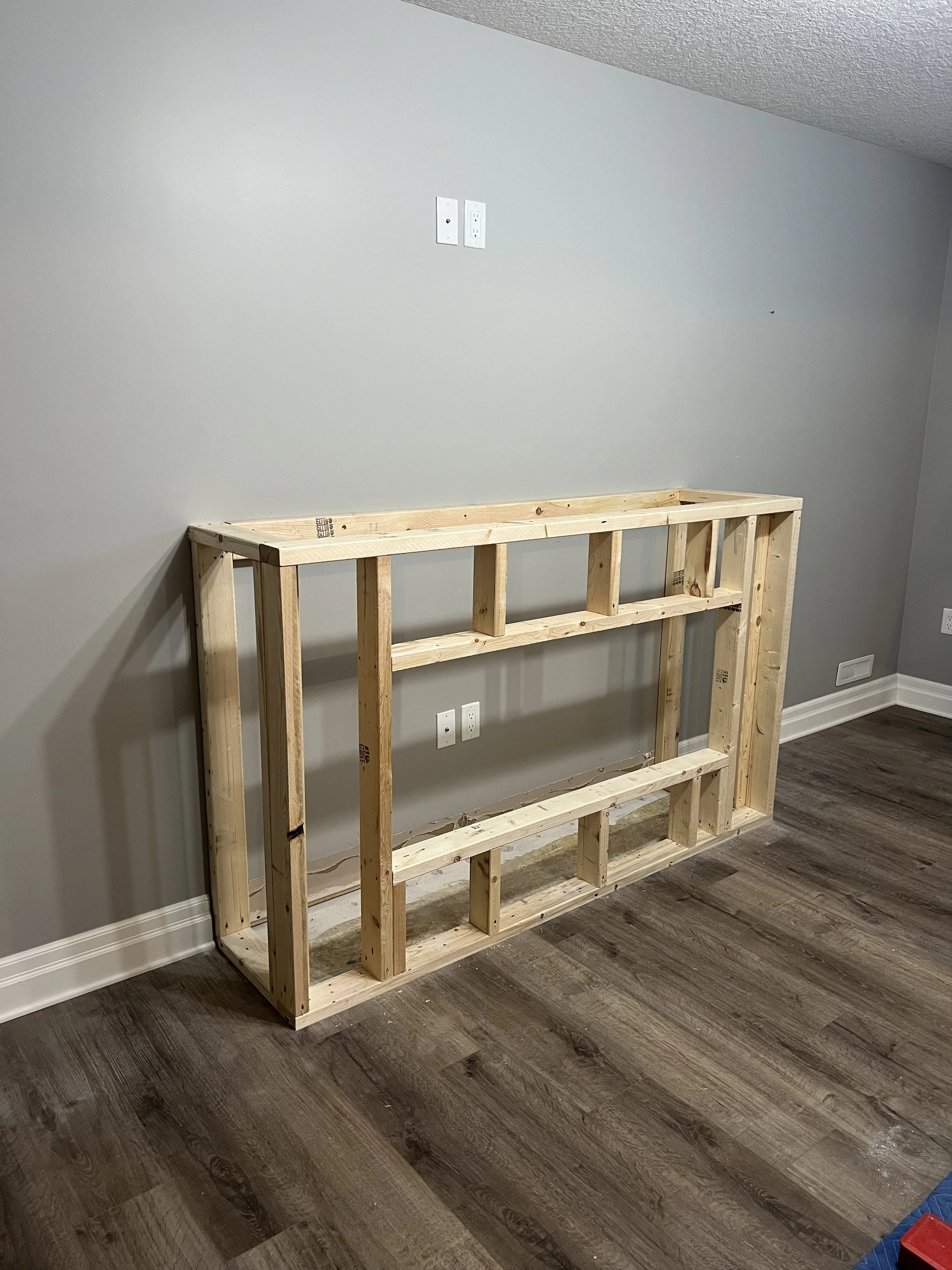 Unfinished wooden frame against a gray wall on a hardwood floor, with electrical outlets visible in a basement renovation.
