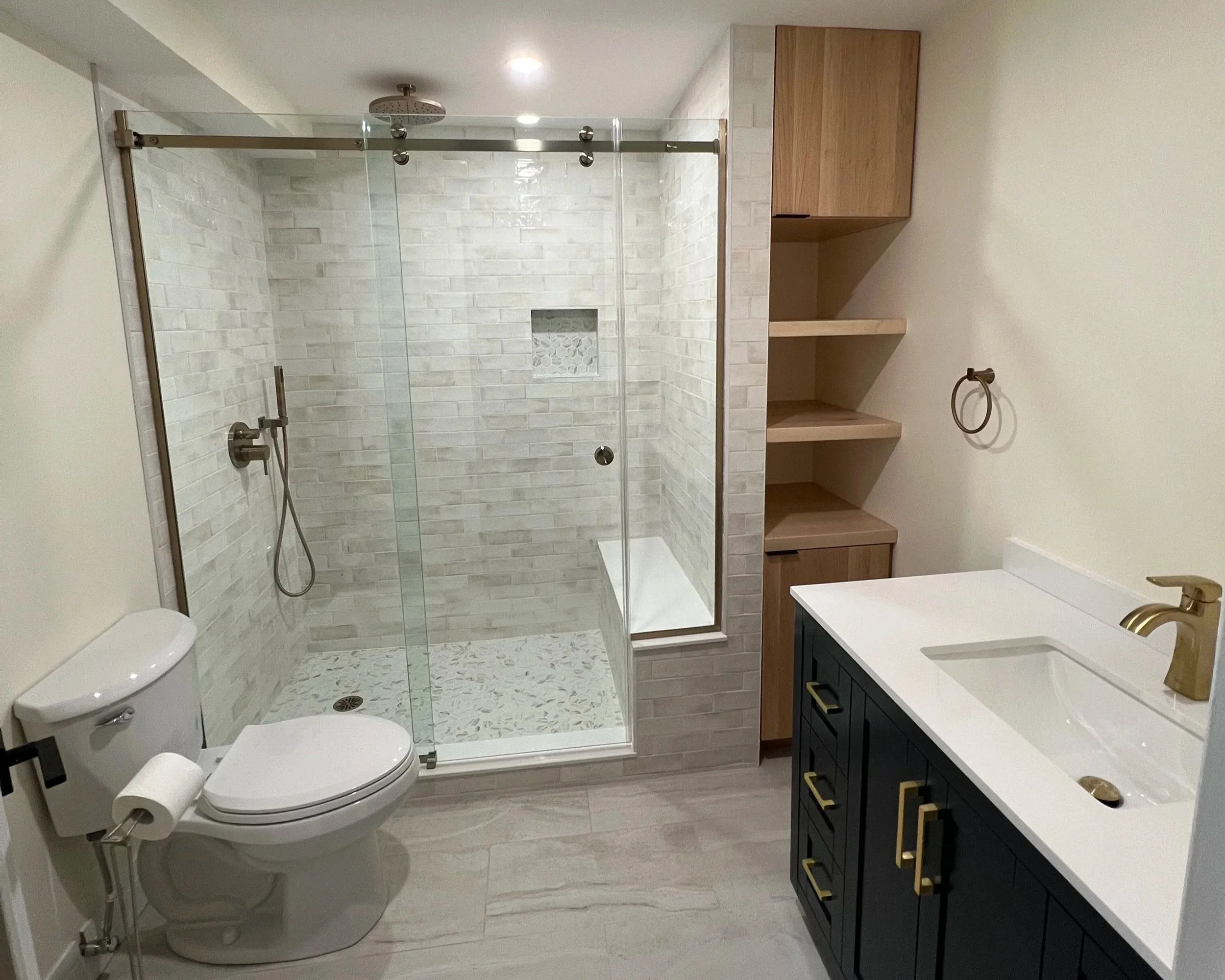 finished bathroom renovation featuring a glass-enclosed walk-in shower with white brick tiles, a built-in shelf unit, a white countertop with an integrated sink, a gold faucet, a wall-mounted towel ring, and wooden open shelves.