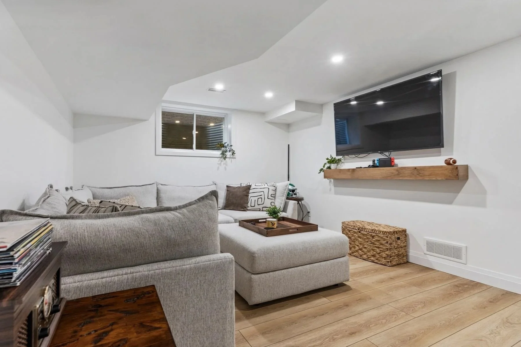 Finished basement with a gray sectional sofa, a wall-mounted flat-screen TV on a white wall, a wooden shelf below the TV, and wooden flooring.