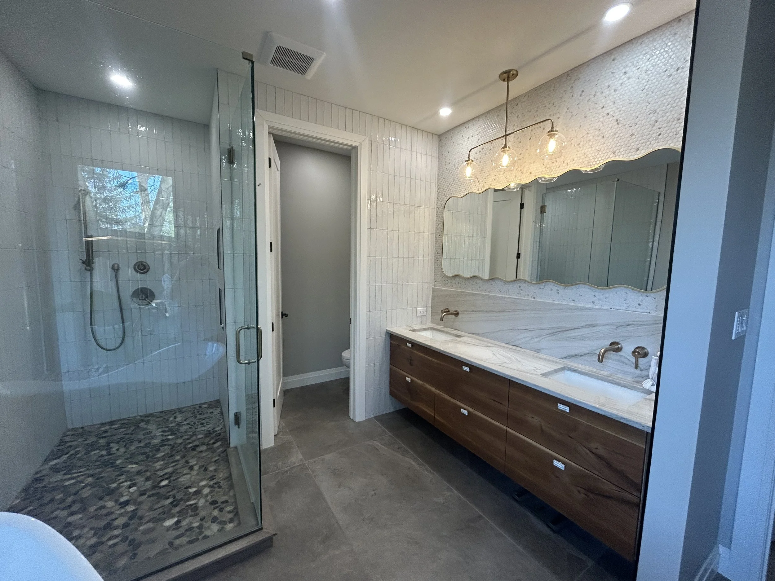 Modern bathroom renovation with a walk-in shower, wooden vanity with a marble countertop, large mirror, and lighting fixtures, with gray tiled floors and white wall tiles.