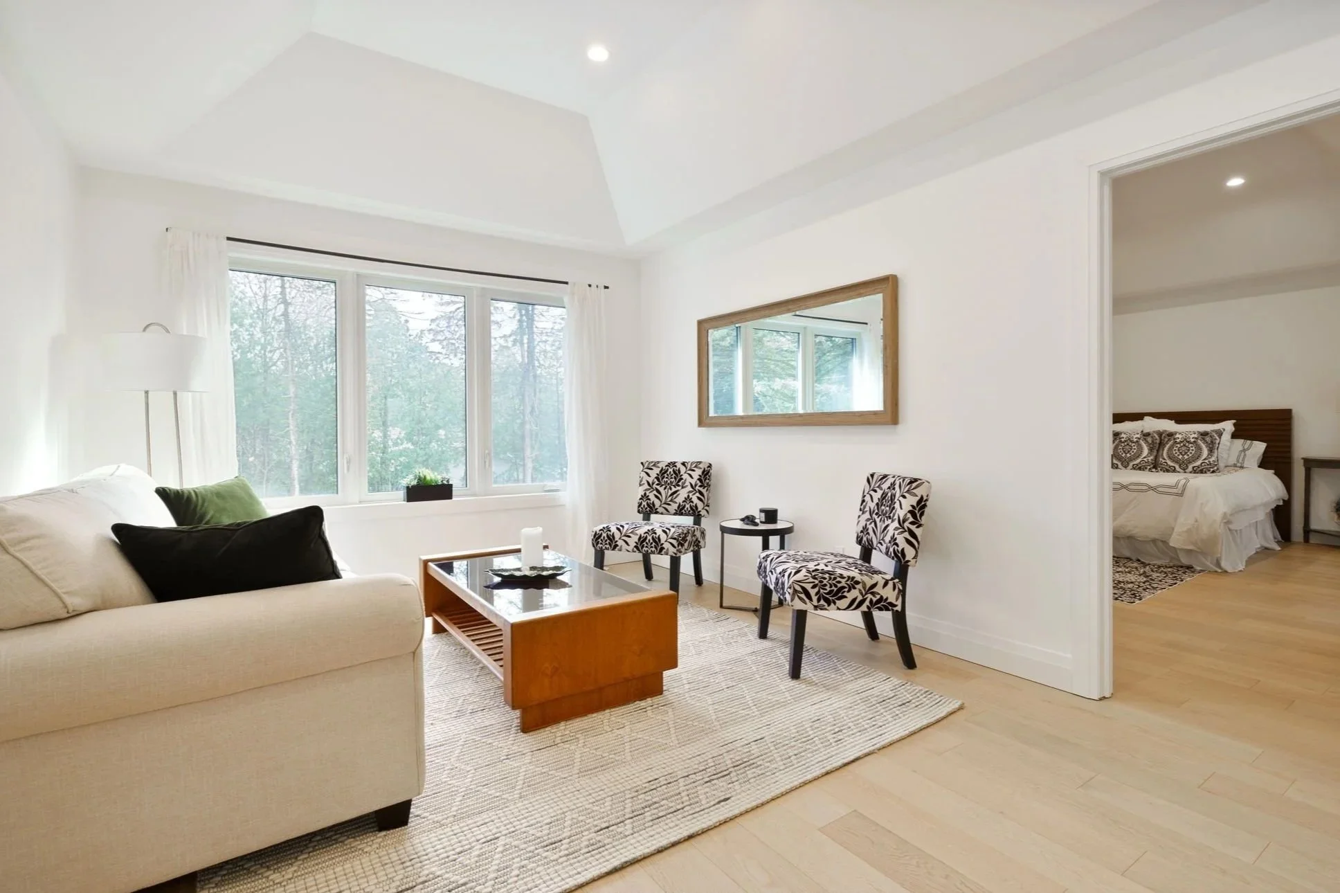 Bright living room with a beige sofa, black and green pillows, a wooden coffee table, two patterned chairs, a large window with white curtains, a mirror on the wall, and a view of a bedroom through an open door.