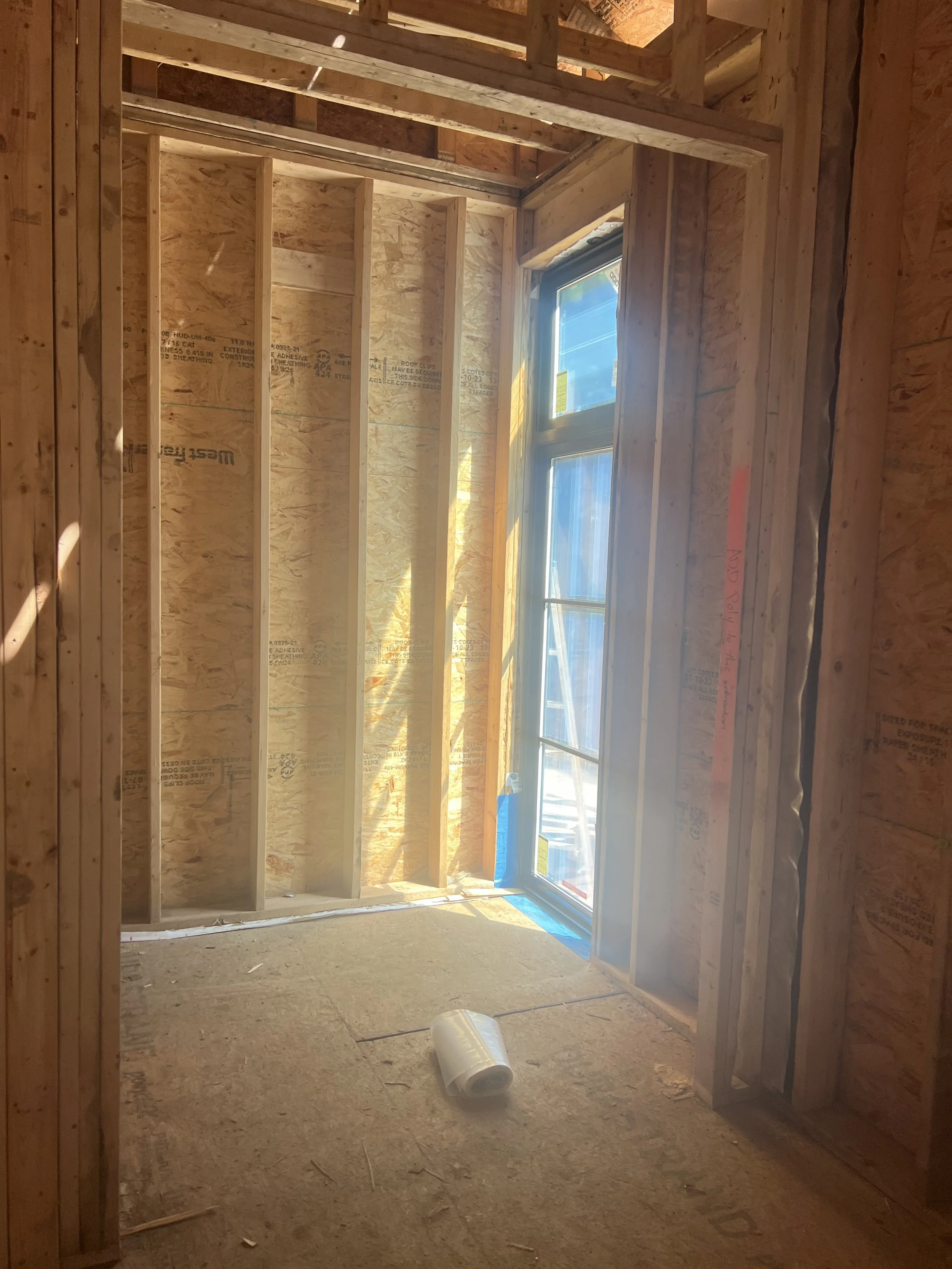 Interior view of a room under construction with wooden framing, exposed plywood walls, a window, and a roll of plastic on the floor.