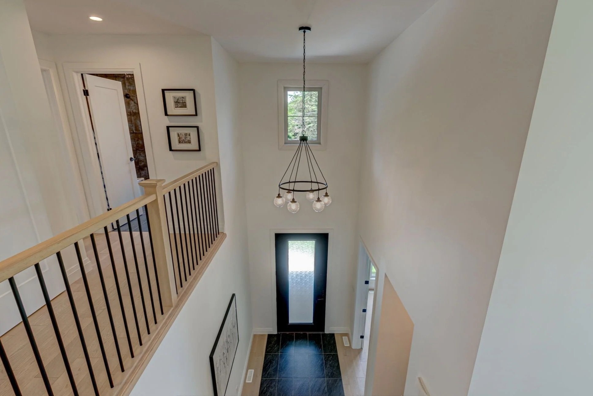 The image shows a view of a modern, two-story home's entryway and upstairs landing area. There is a black front door with a frosted glass panel at the bottom, and a small window above it. A contemporary chandelier with exposed bulbs hangs from the ceiling. The upstairs area features a wooden railing with black vertical balusters. The walls are painted white, and there is a narrow window on the upper level, allowing natural light through. Decorative framed pictures are on the wall near a doorway to a bathroom, which can be seen with tiled walls.