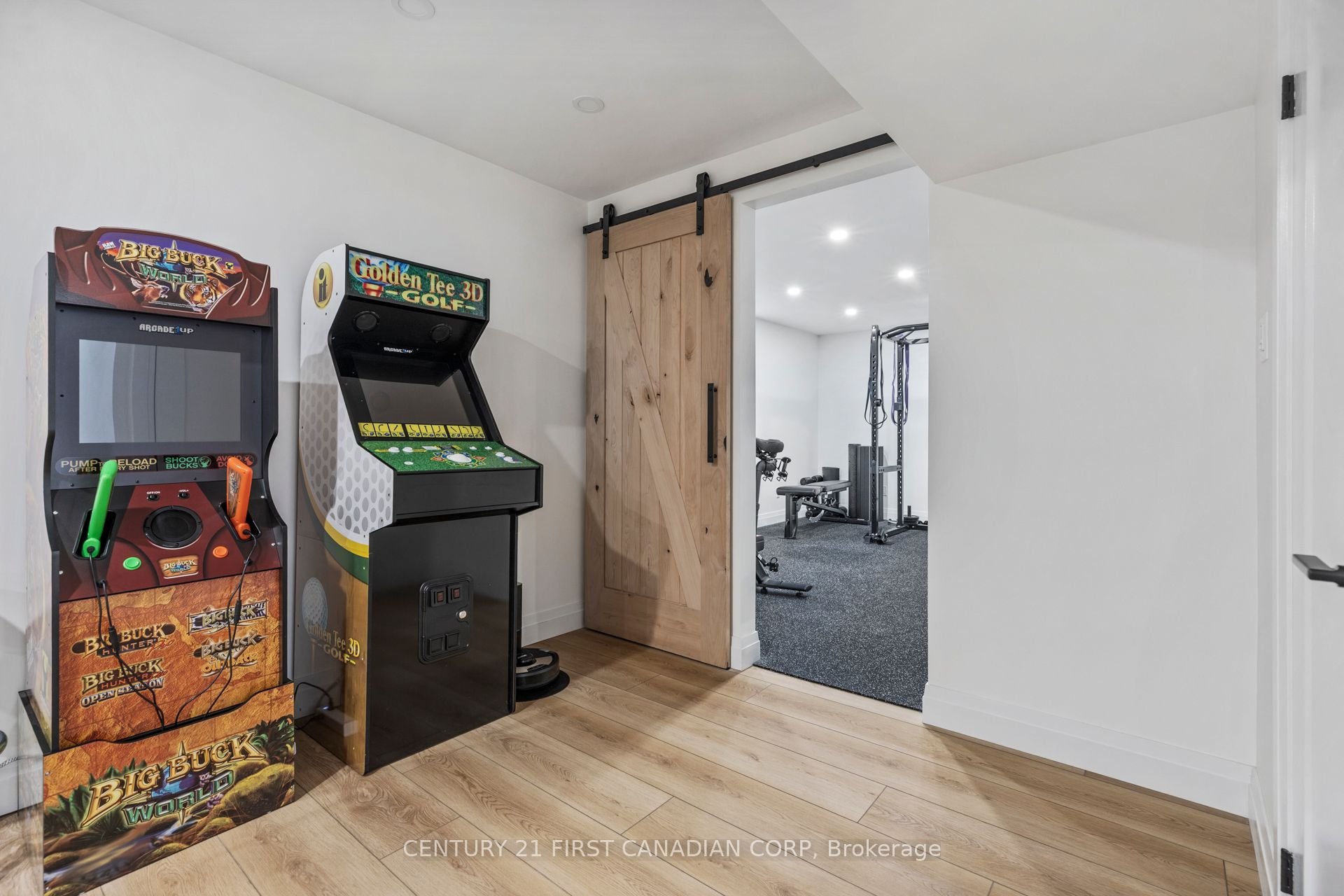 Basement renovation with an arcade game machines including Big Buck Hunter and Golden Tee 3D Golf in a home game room, with a door leading to a gym area in the background.