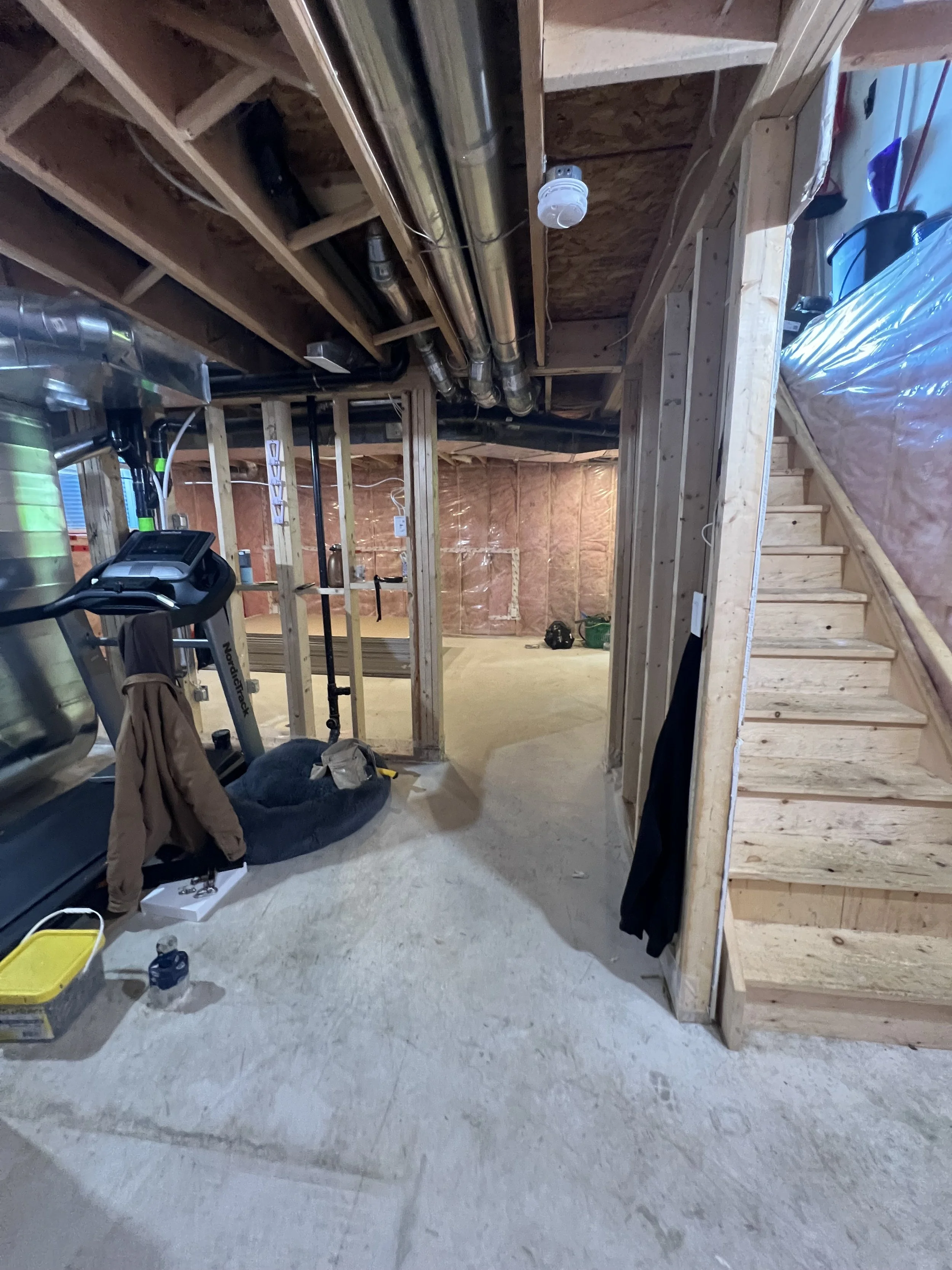Interior of a basement renovation under construction with exposed wooden framing, stairs on the right, plumbing pipes on the ceiling, and various construction tools and materials on the floor.