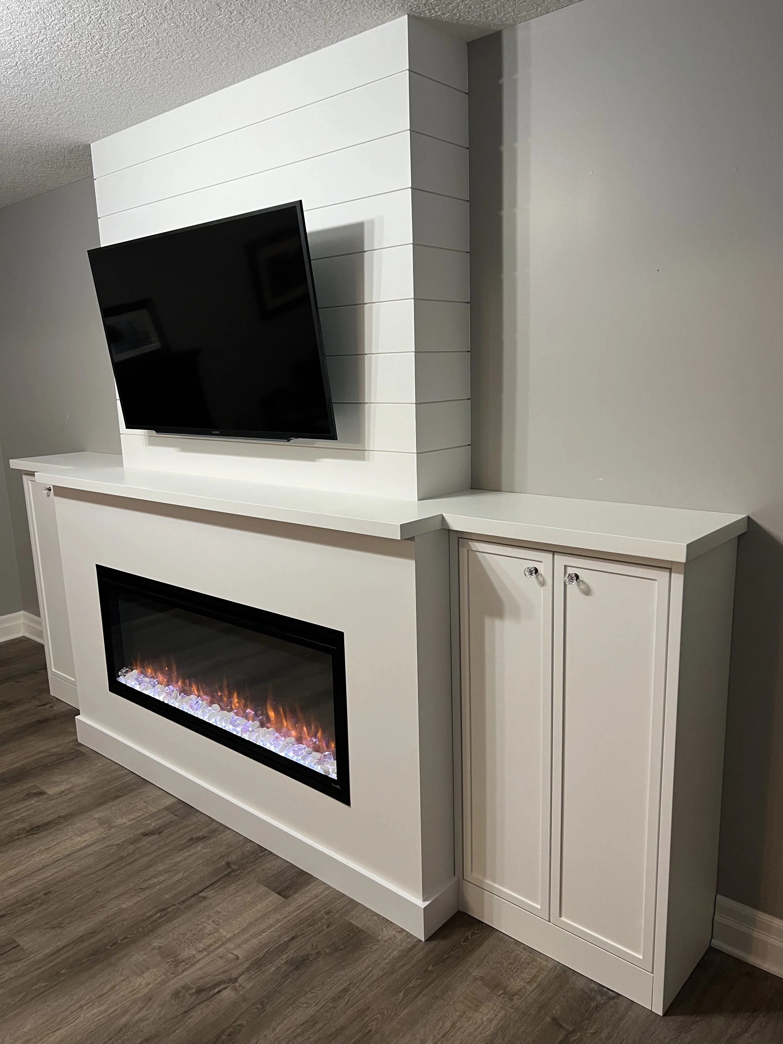 Basement renovation entertainment unit with a wall-mounted flat-screen TV above an electric fireplace, with a white cabinet on the side.