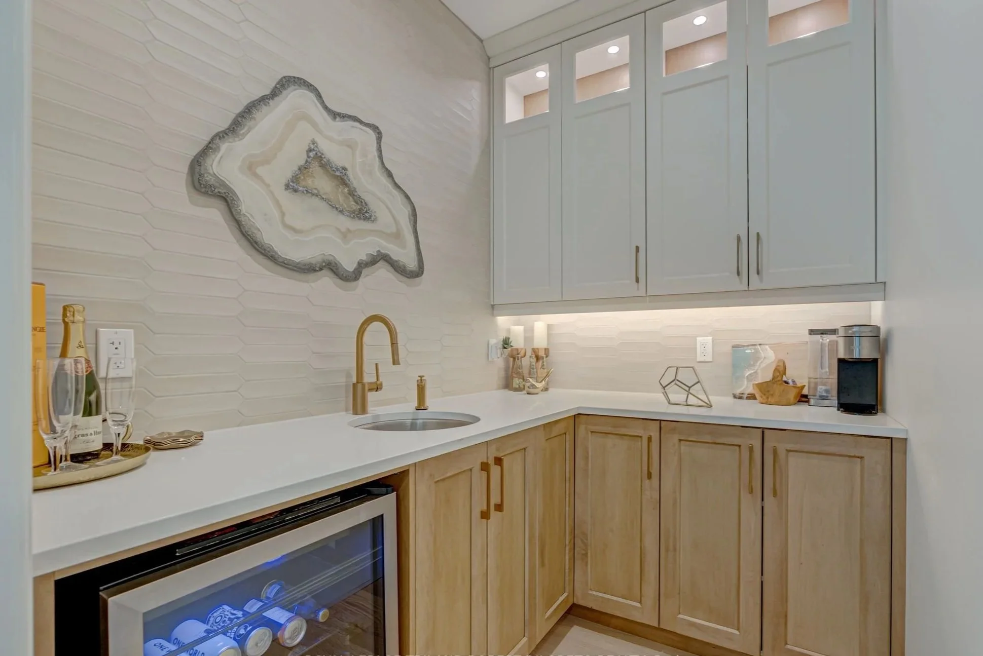 A kitchen pantry renovation with a white countertop, light wooden lower cabinets, and white upper cabinets. There's a decorative stone-like wall art, a gold faucet, and a mini wine cooler with drinks inside. 