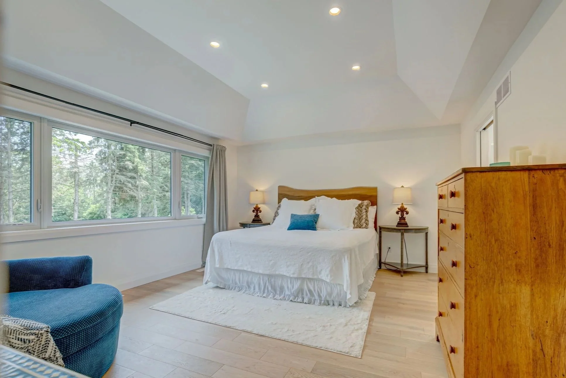 Bright bedroom with large window, white bedding, wooden furniture, and green trees outside.