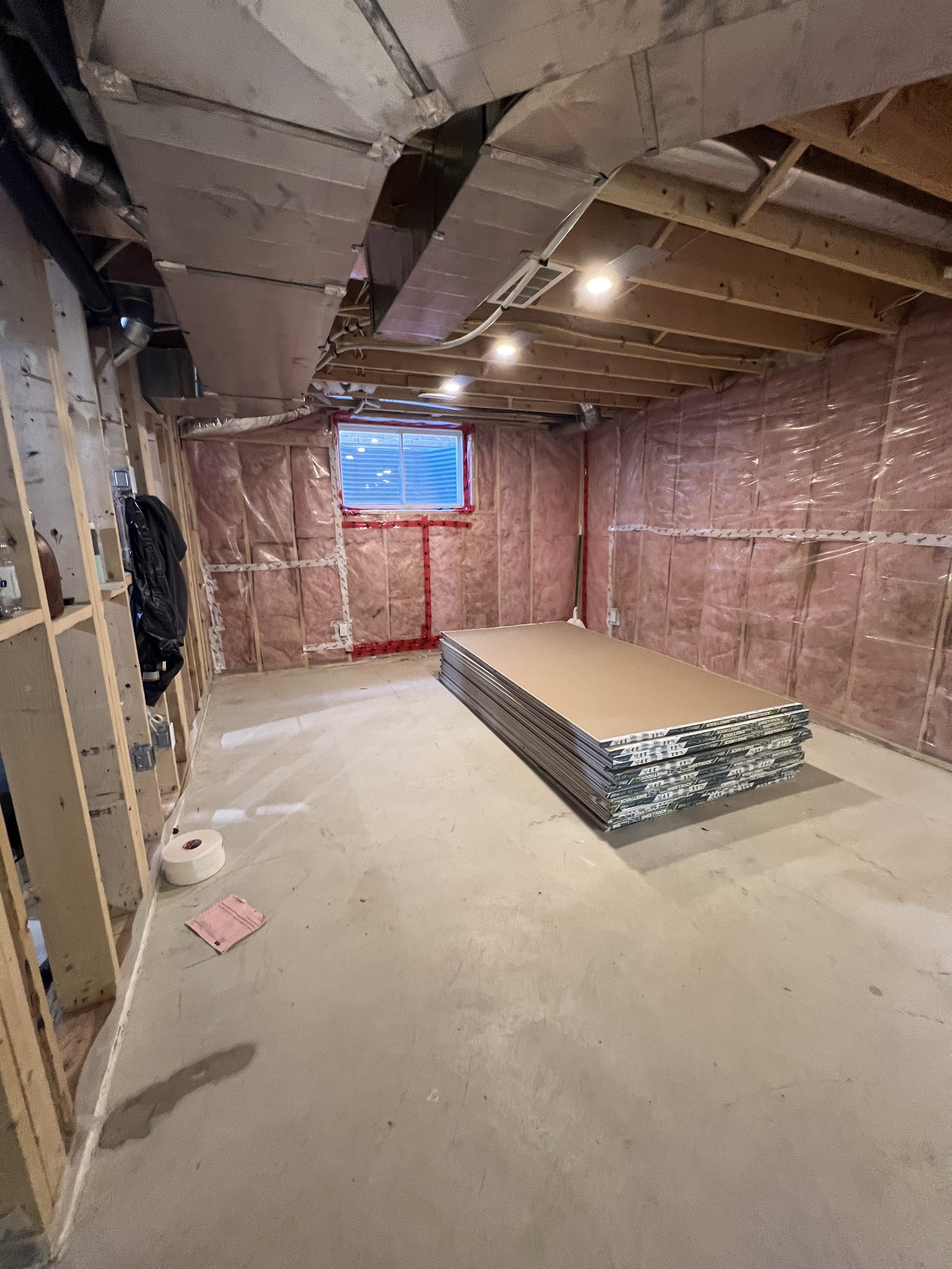 Basement renovation with exposed wooden framing, insulation on the walls, a egress window, and stacked drywall sheets on the floor.