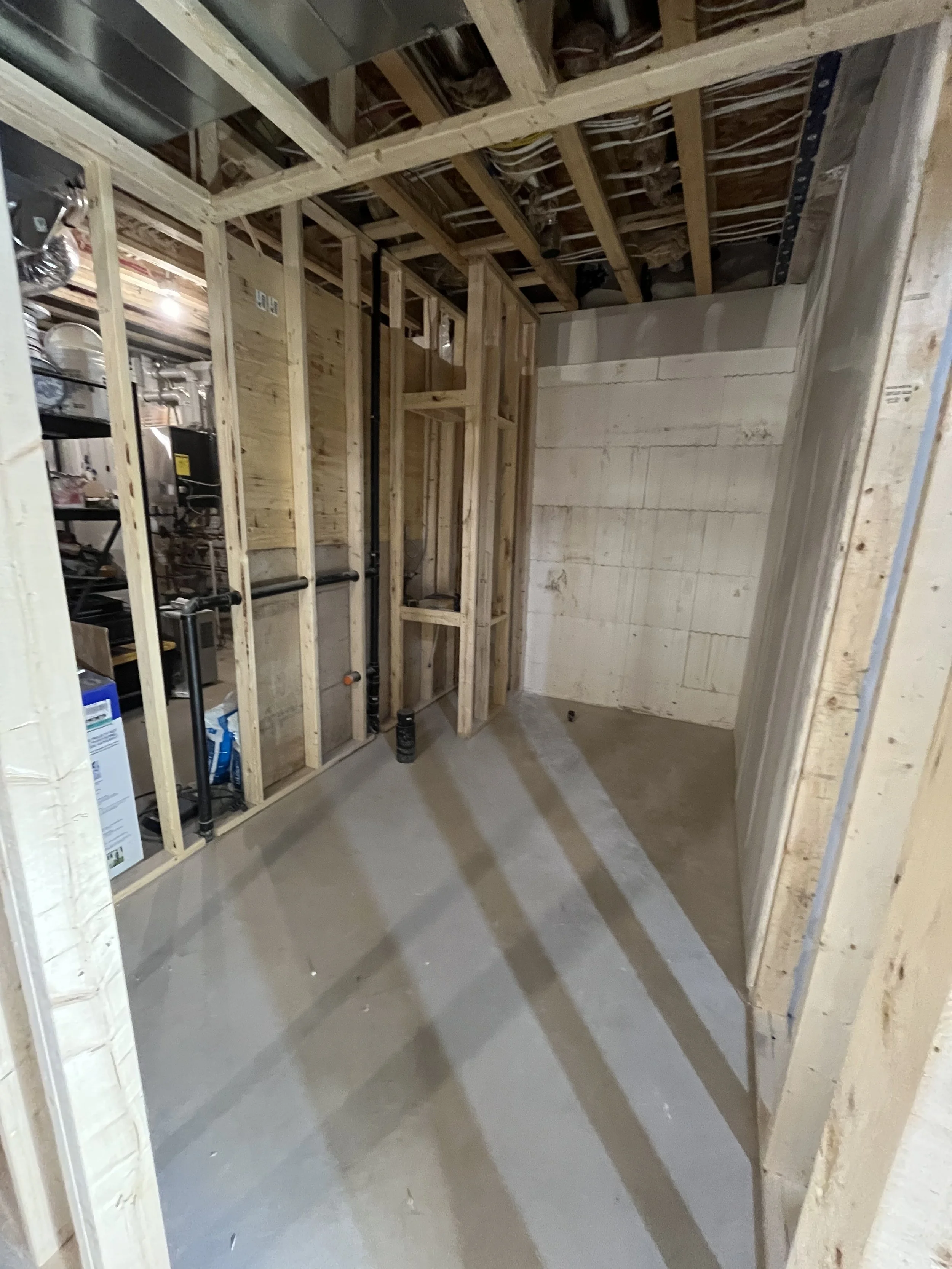 An unfinished basement renovation under construction with exposed wooden framing, a concrete floor, and some piping installed along one wall.