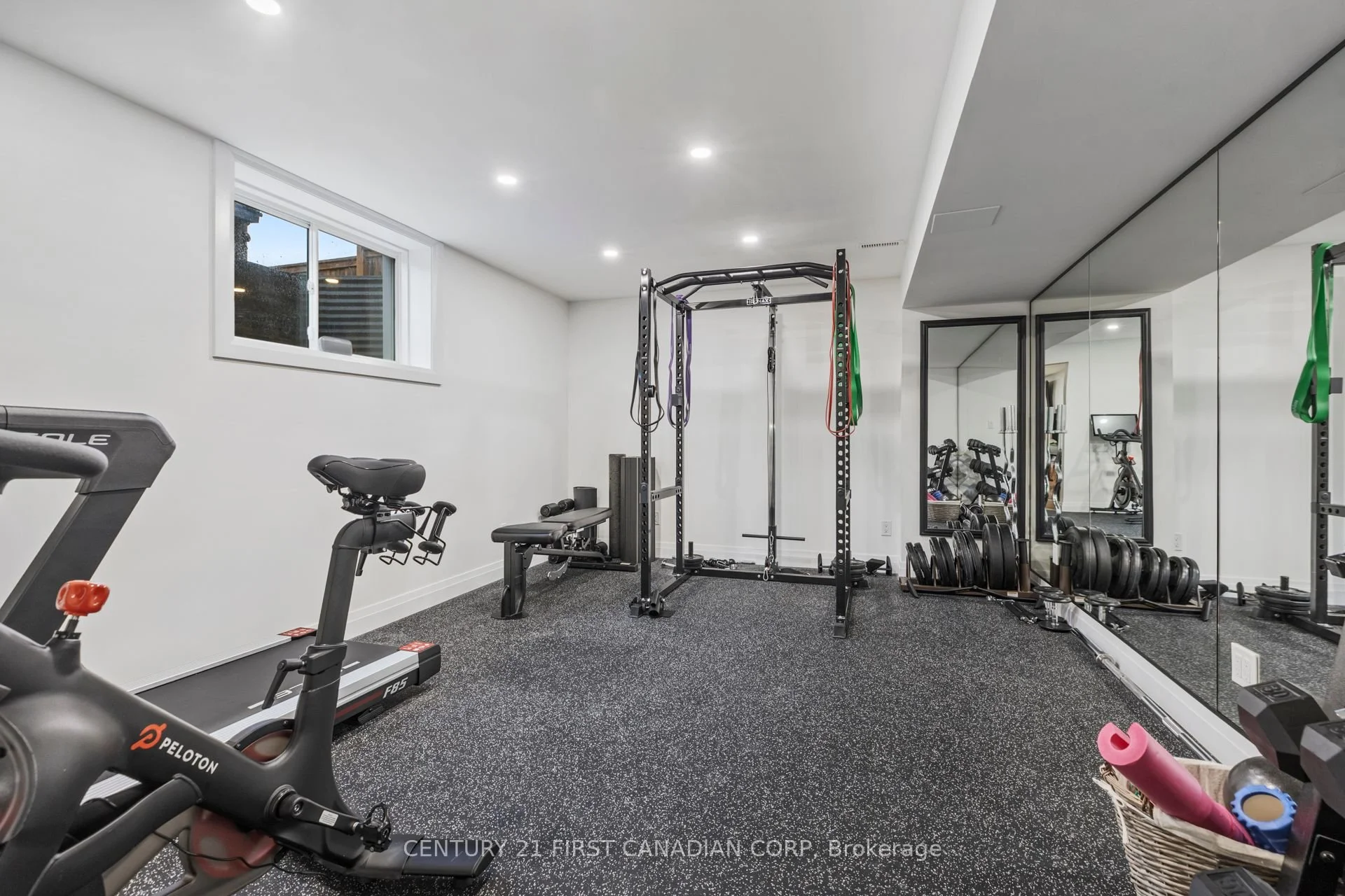 Home gym in finished basement renovation with Peloton exercise bike, weight bench, pull-up bar, free weights, resistance bands, and large mirror on the wall.