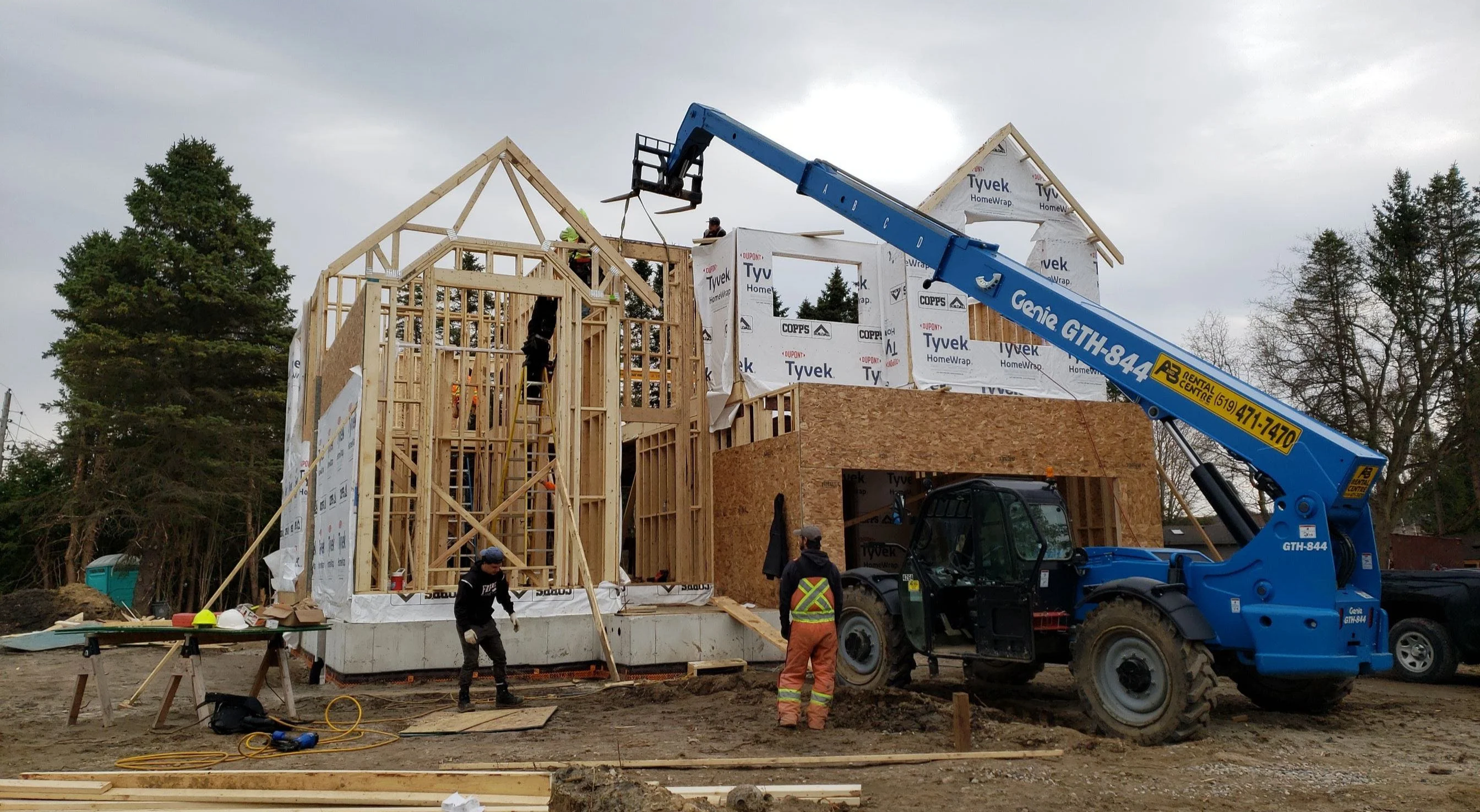 Construction site with workers building a house, using a large blue crane to lift and position wooden framing. The house is in early framing stages, with some walls covered in Tyvek house wrap. Surrounding trees and overcast sky are visible.