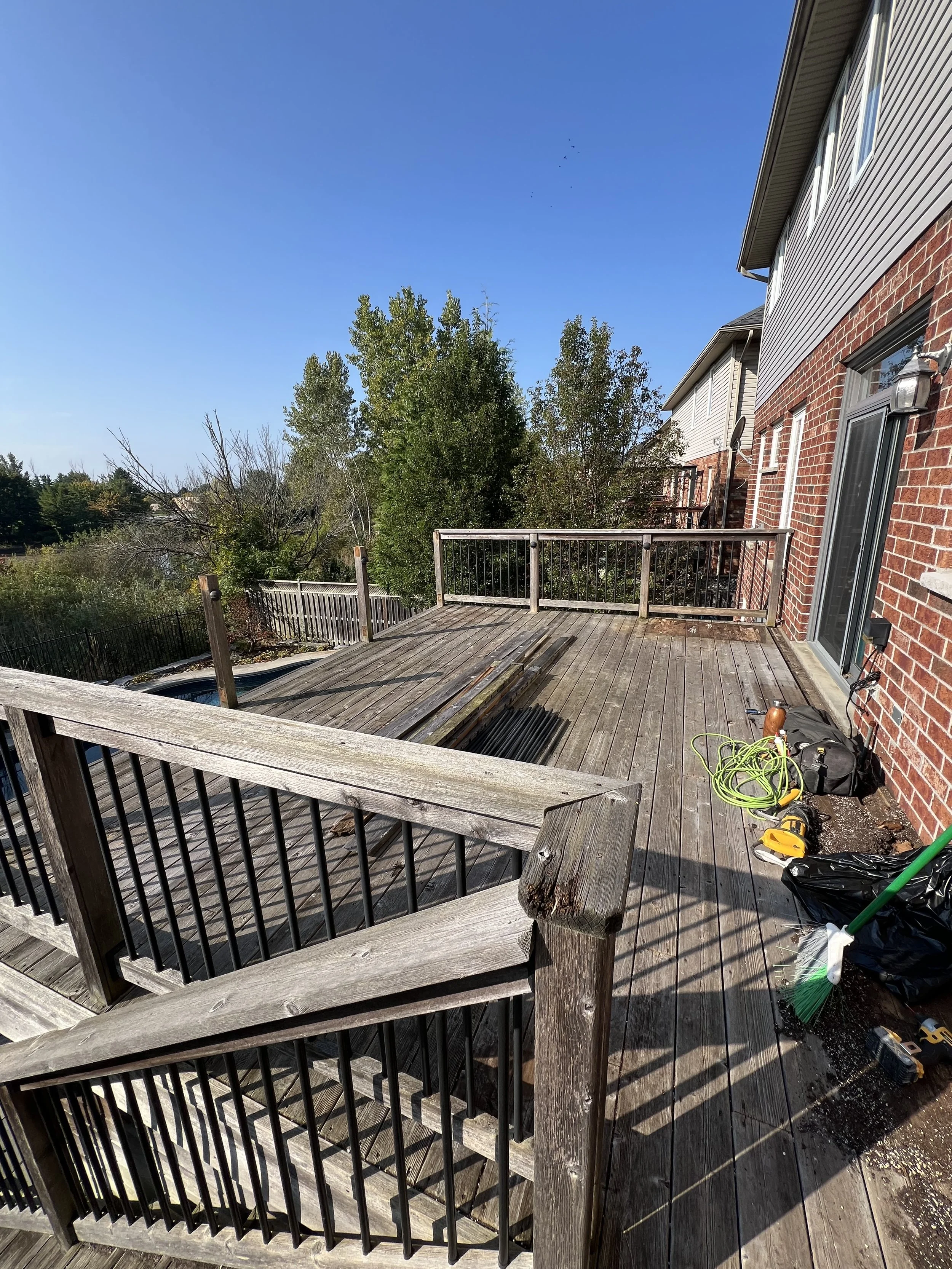 An outdoor wooden deck attached to a brick house, with gaps in the wood planks, some construction tools, a broom, a bag, and a coiled extension cord. The deck has railings and is surrounded by trees and greenery, with a clear blue sky overhead.