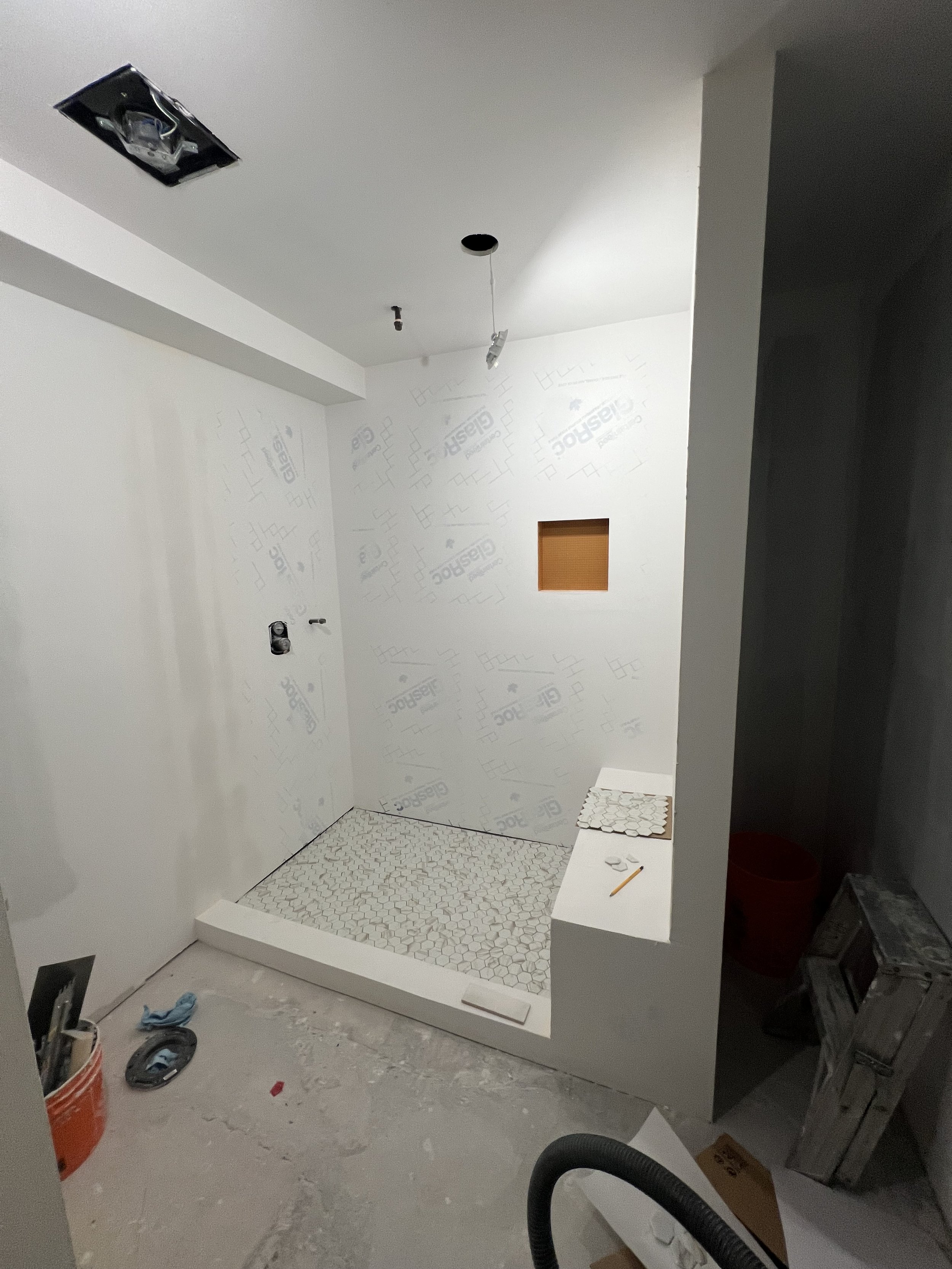 Under construction bathroom with tile shower area, unfinished walls, and construction materials and tools on the floor and ledge.