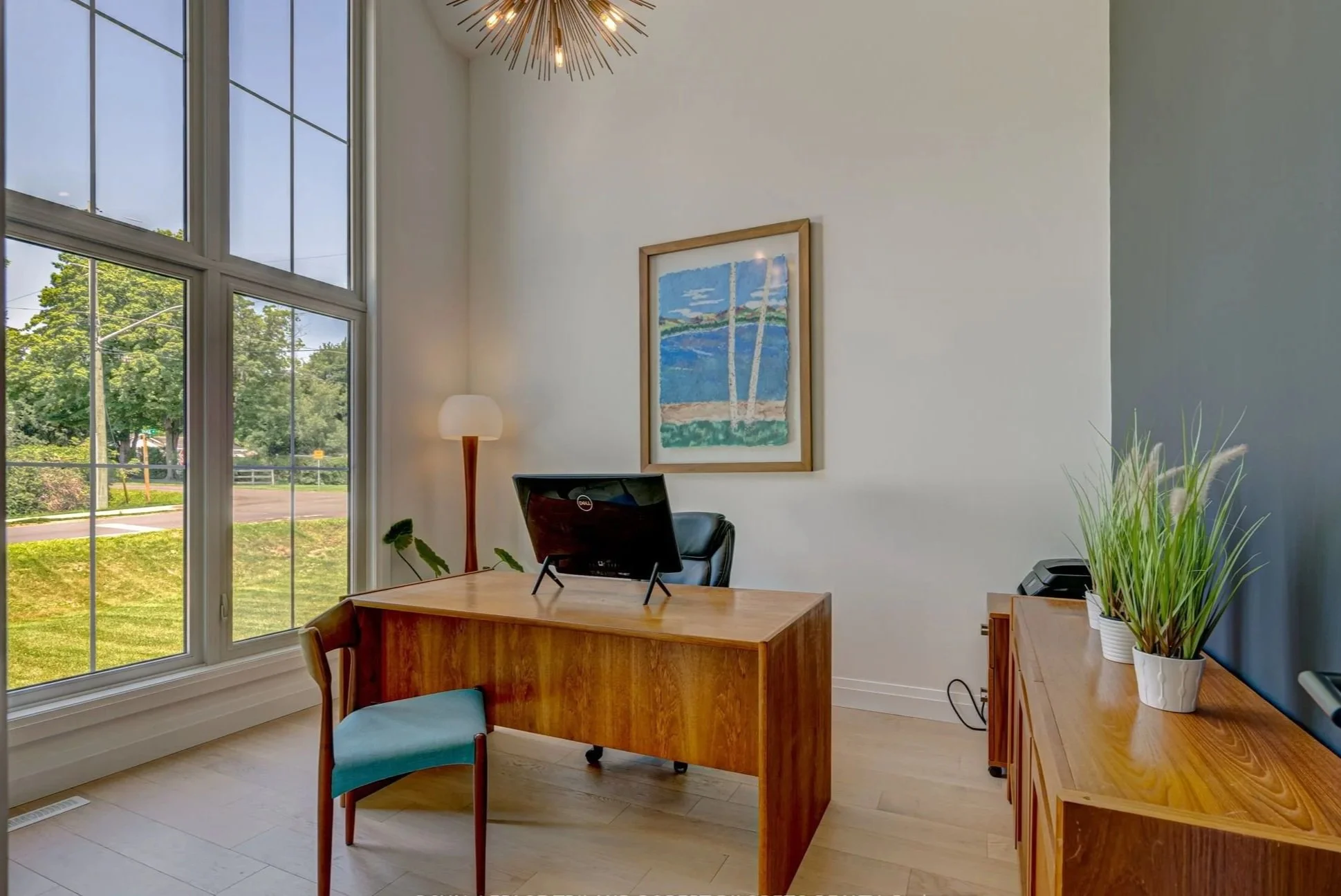 An office with a large window showing trees outside, a wooden desk with a computer monitor, and a wooden sideboard with potted plants, a painting of a beach scene on the wall, and a standing lamp.
