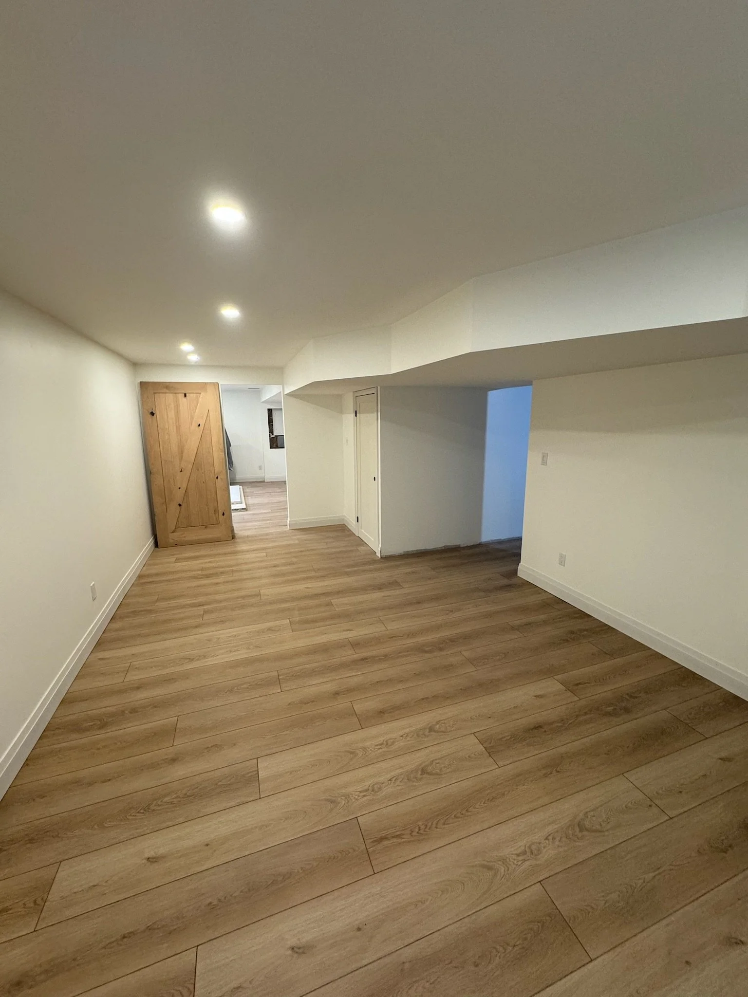Finished basement renovation with wood flooring, white walls, and ceiling with recessed lighting. There is a wooden sliding door on one side and a small alcove or passageway.