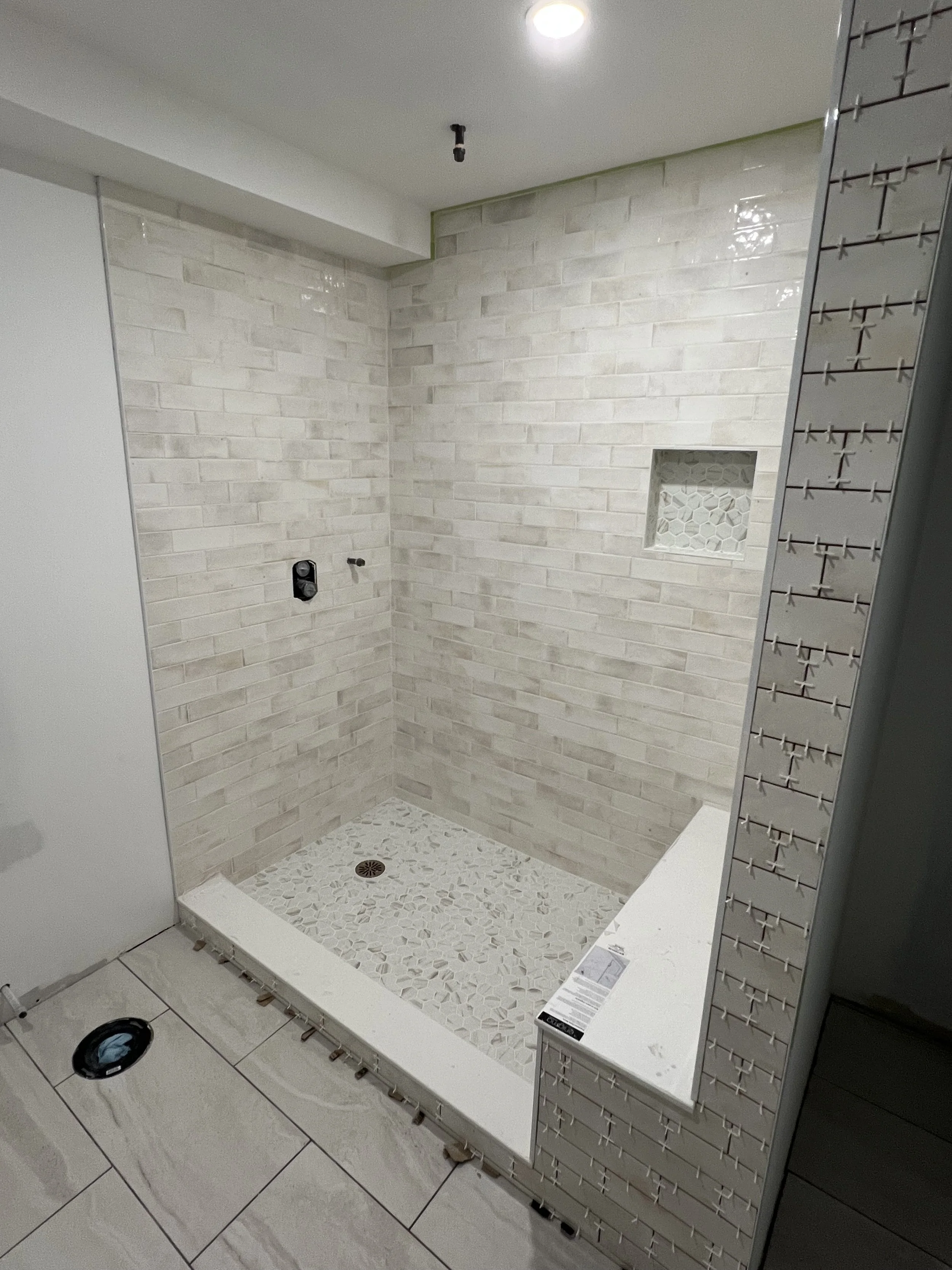 A bathroom shower renovation with beige brick tiles on the walls, a small built-in niche with patterned tiles, and a mosaic tile floor. The shower area is framed by a white ledge, with plumbing fixtures visible but not installed.