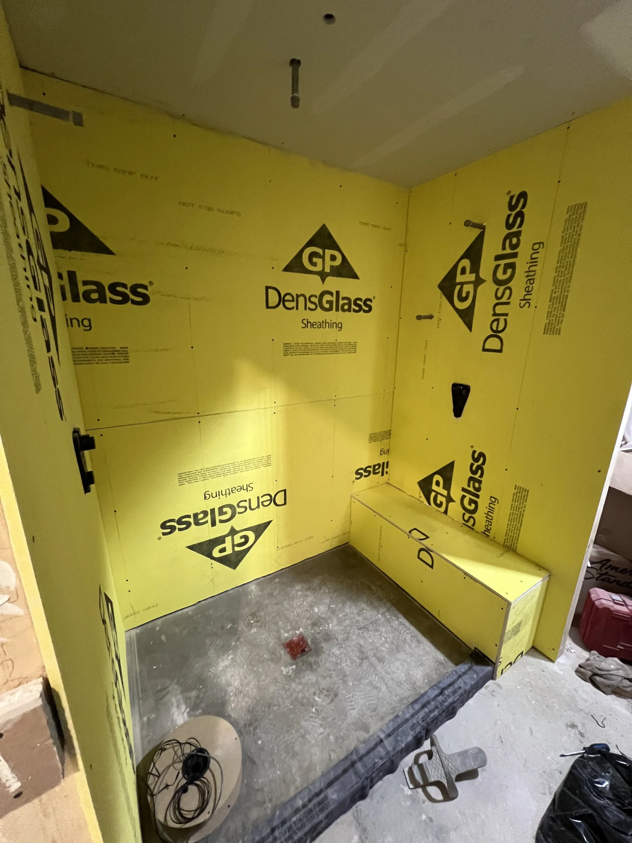 Interior of a bathroom renovation with yellow DensGlass sheathing on the walls, an electrical box on the right wall, and a concrete floor with a red marker and some electrical wires on the left. The ceiling has a light fixture and an uncovered pipe.