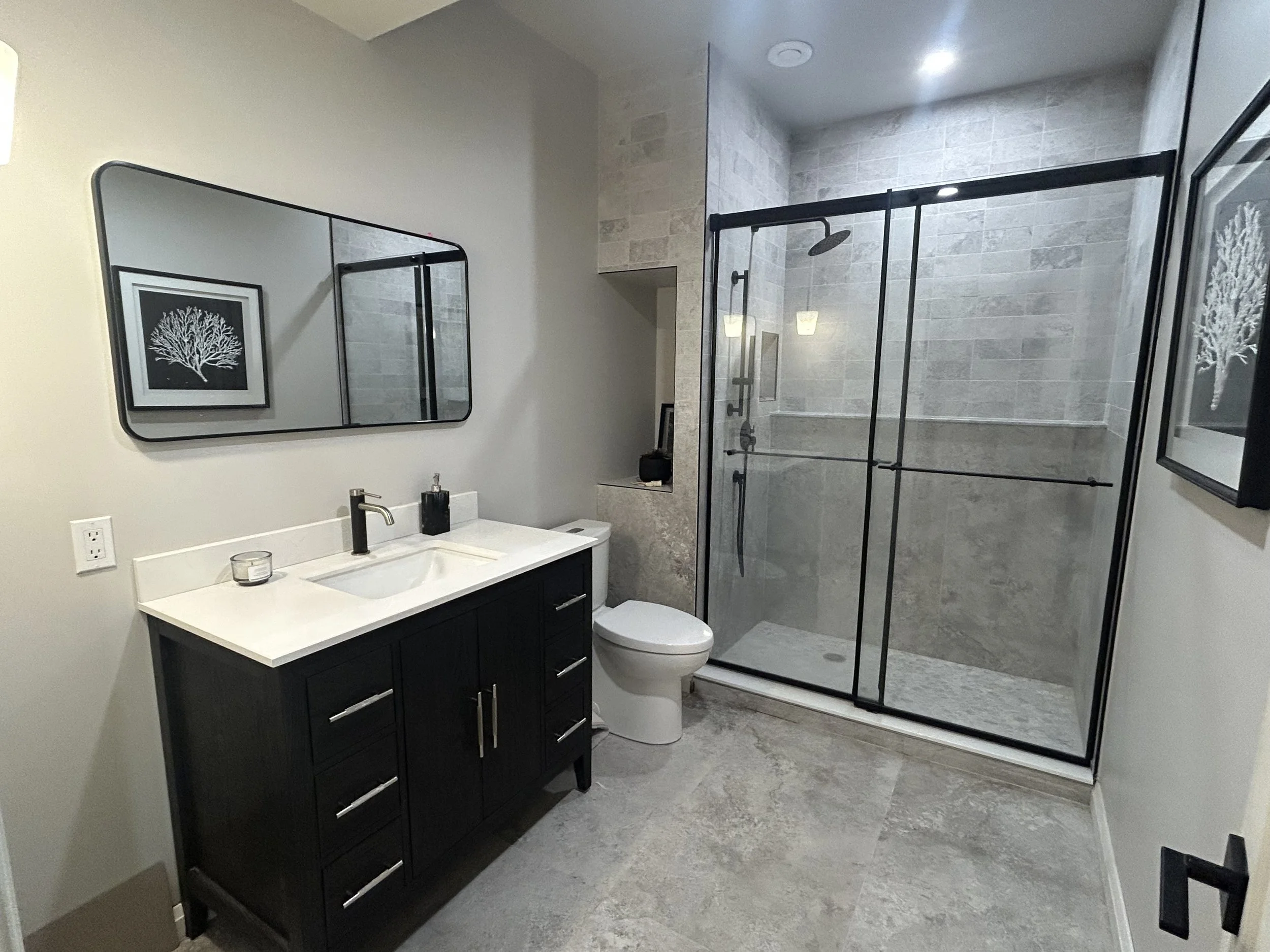 Modern bathroom featuring a black vanity with a white countertop, a mirror above the sink, a framed black and white artwork, a glass-enclosed shower with black framing, and neutral-toned tiles.
