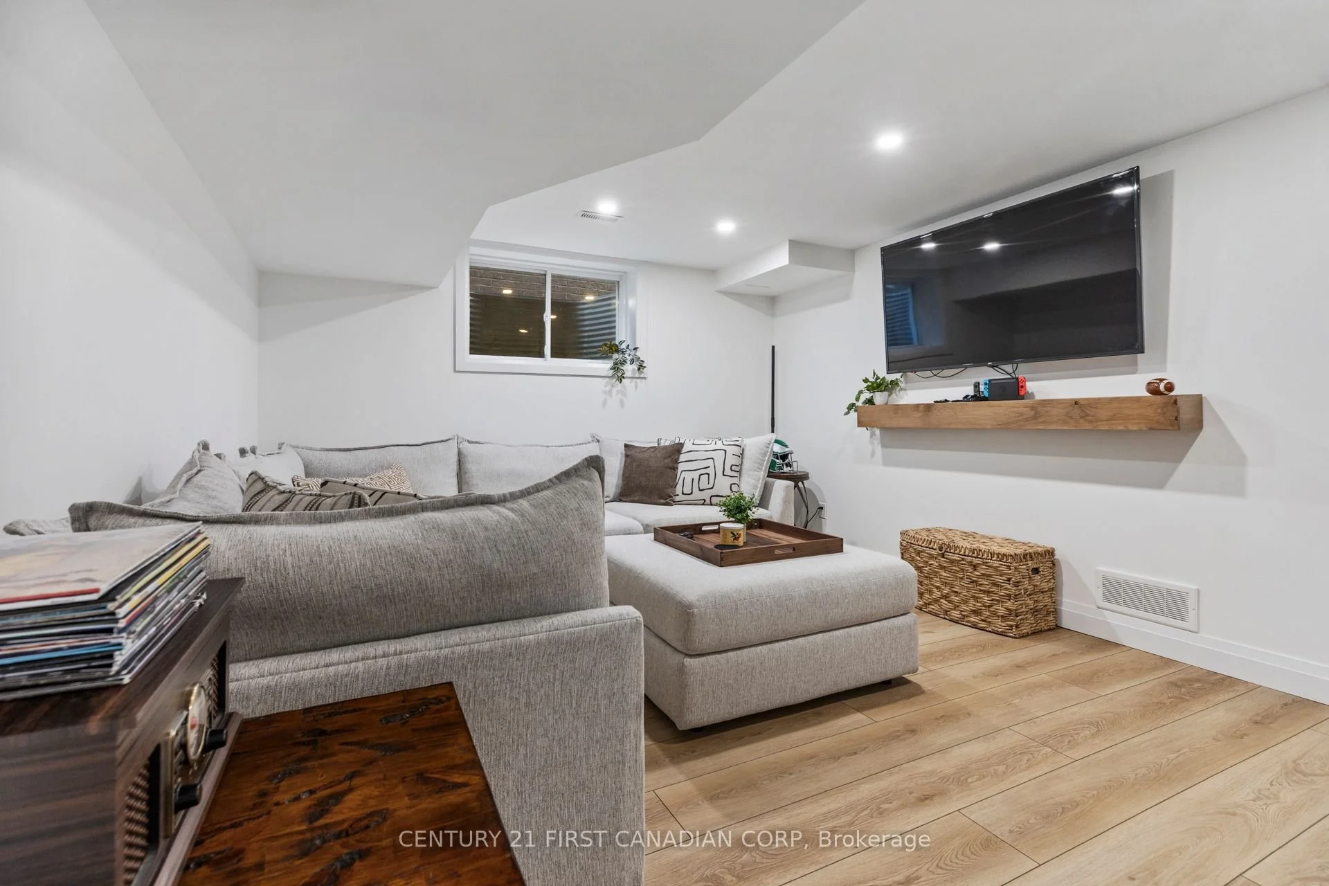 Finished basement renovation with a gray sectional sofa, a wooden coffee table tray with a small plant, a wall-mounted TV on a wooden shelf, a small egress window, and wood flooring.