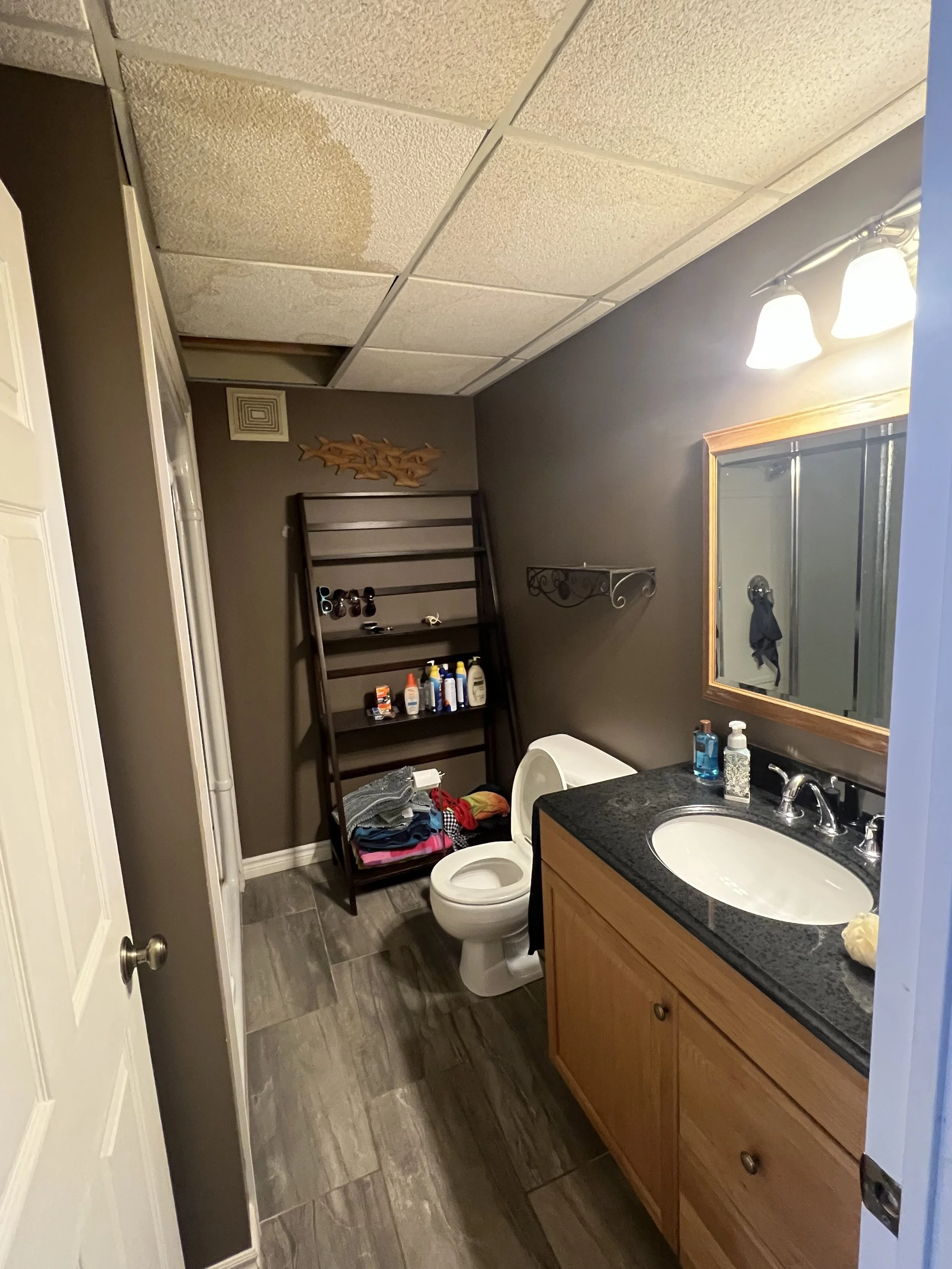 A bathroom with a dirt-stained ceiling, gray walls, a wooden vanity with a black countertop, a mirror, and a toilet. There is a black metal shelf with toiletries and clothing, and a mirror with light fixtures above the sink.