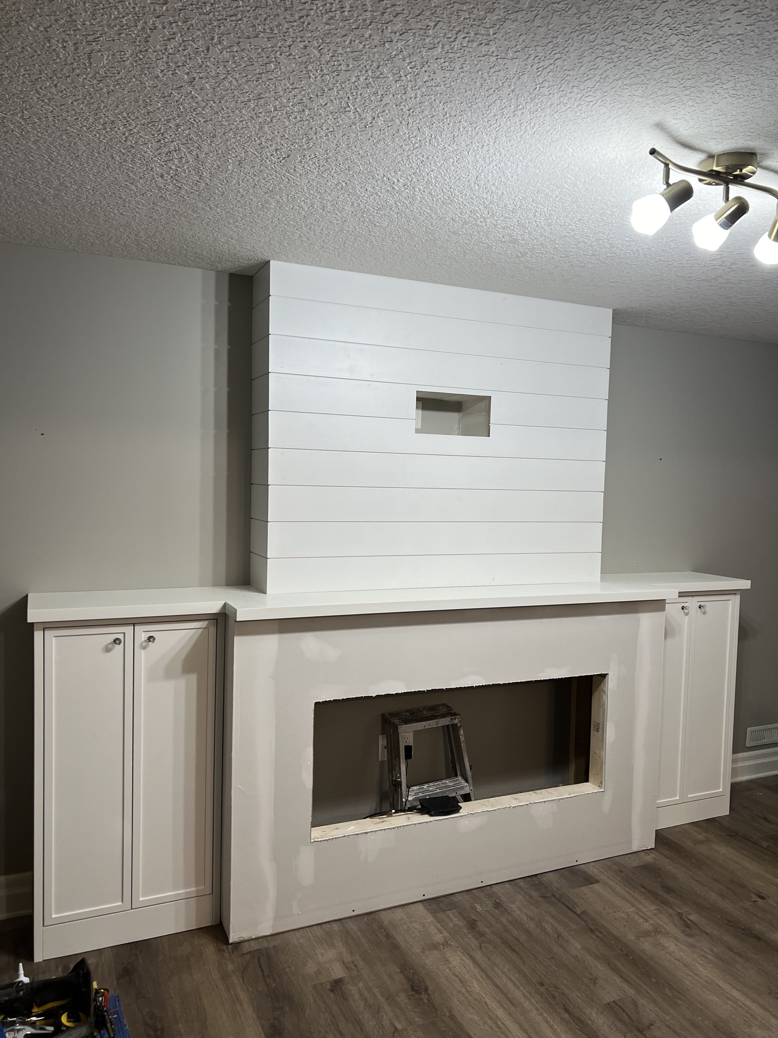 Basement renovation with a fireplace under renovation with white built-in cabinets on both sides, a framed opening for the fireplace, and a ladder inside the opening. Ceiling with a track light fixture and wood flooring.