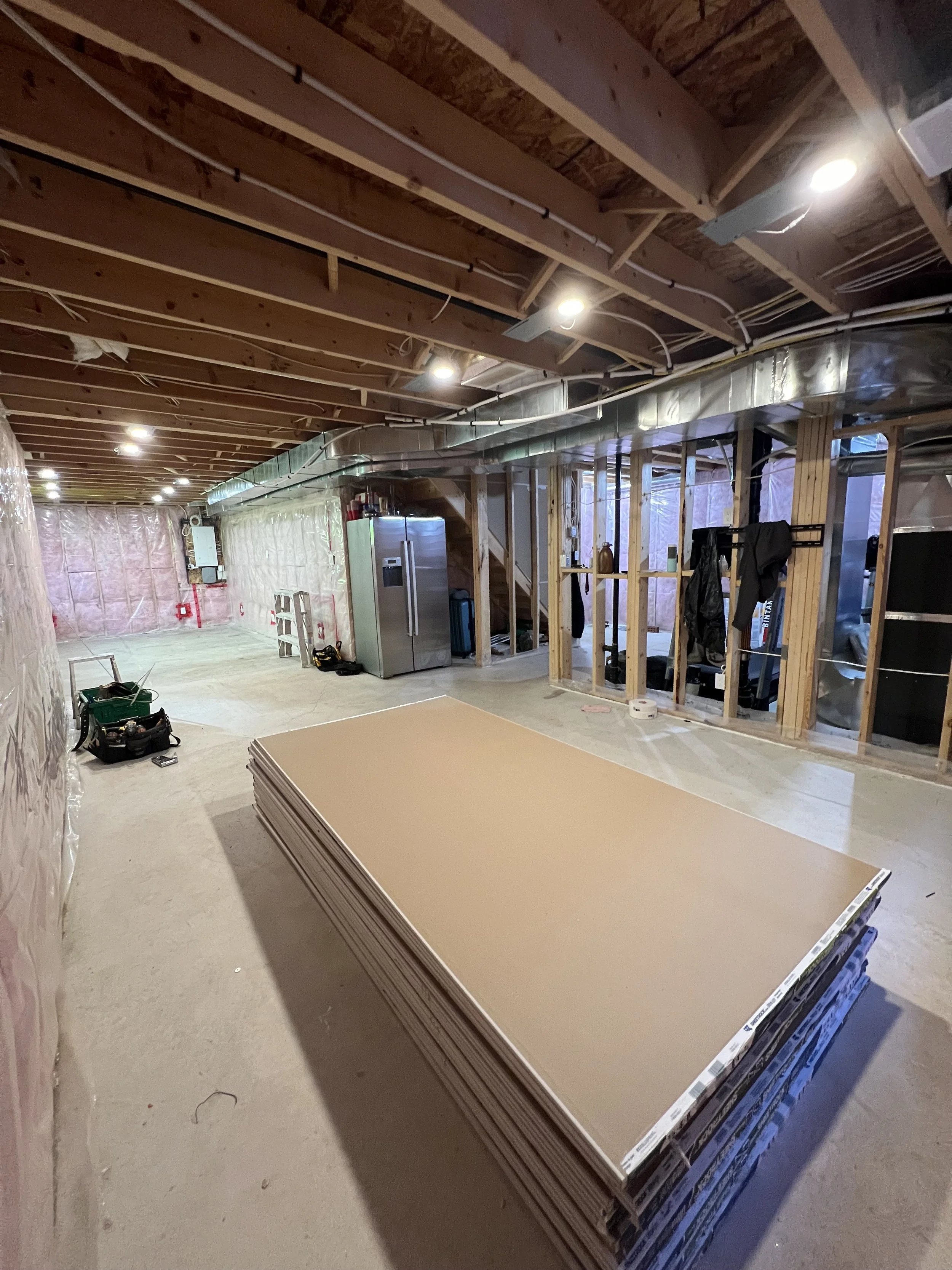 Basement renovation with unfinished walls and ceiling, construction materials, and tools, including a stack of drywall sheets and a refrigerator.