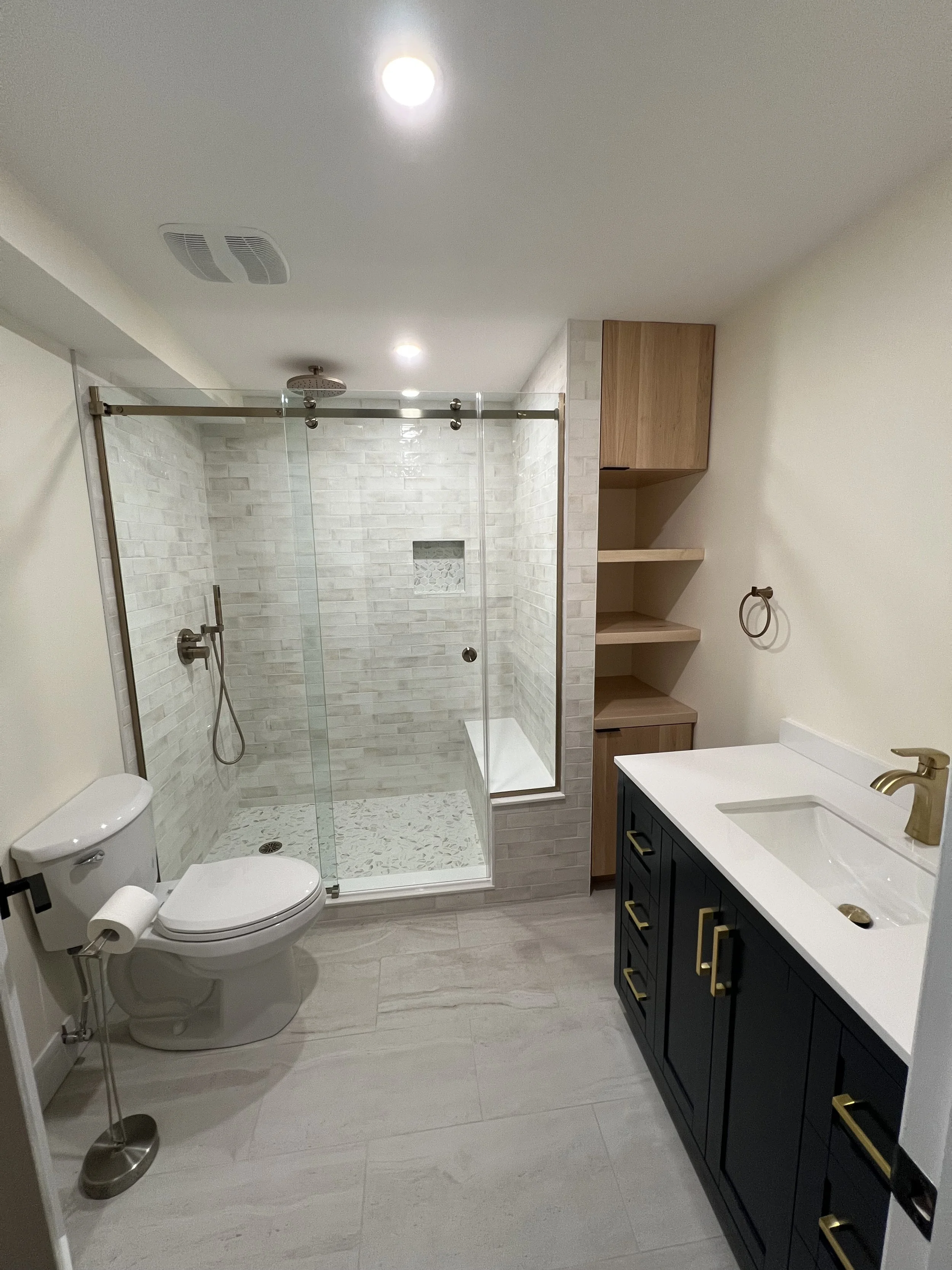 Modern bathroom with a walk-in shower, toilet, dark blue vanity with gold handles, white countertop and sink, brass faucet, wooden shelves, and beige tile flooring.