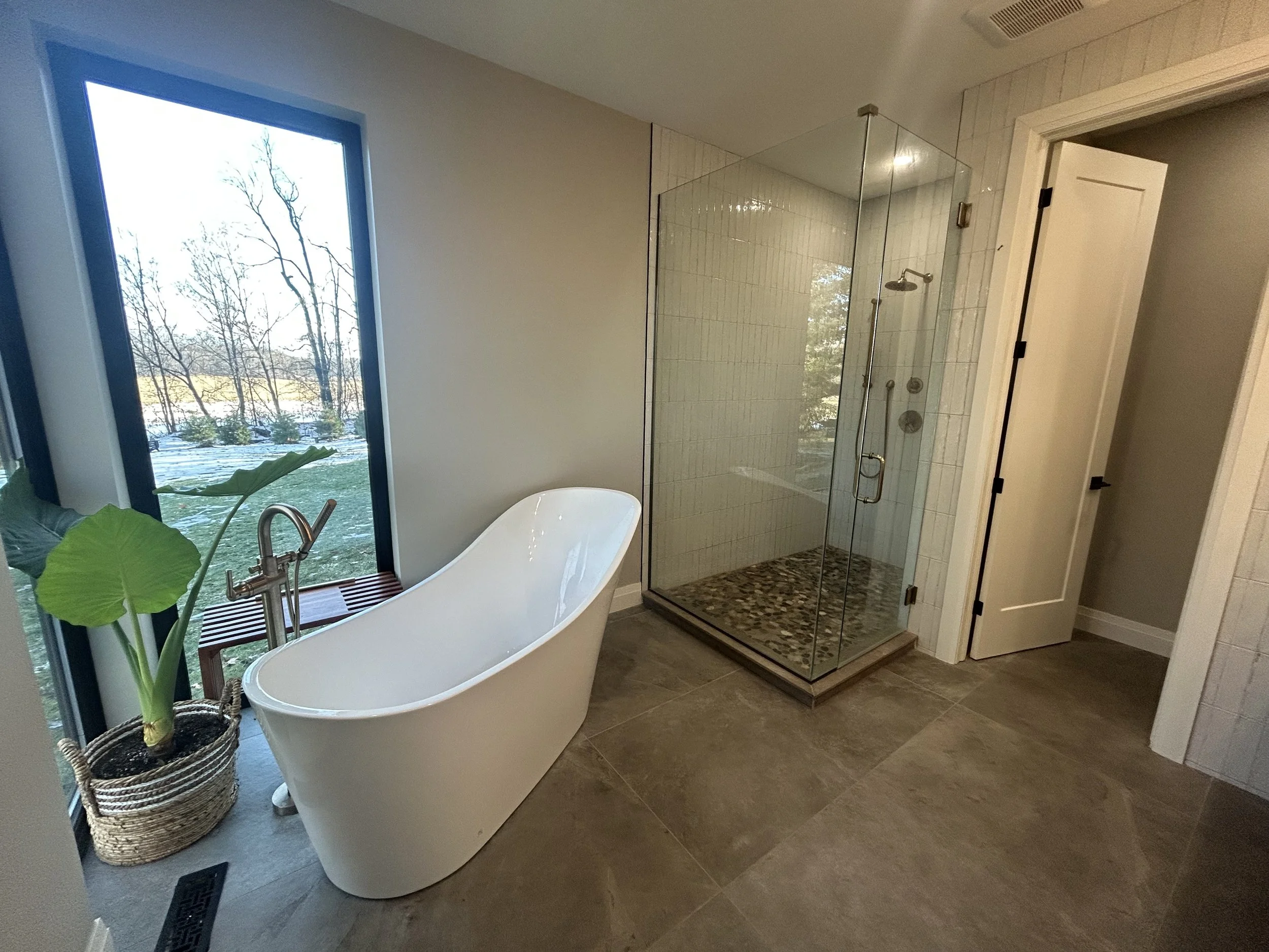 Modern bathroom with large window, standalone bathtub, potted plant, and glass-enclosed shower with pebble floor.
