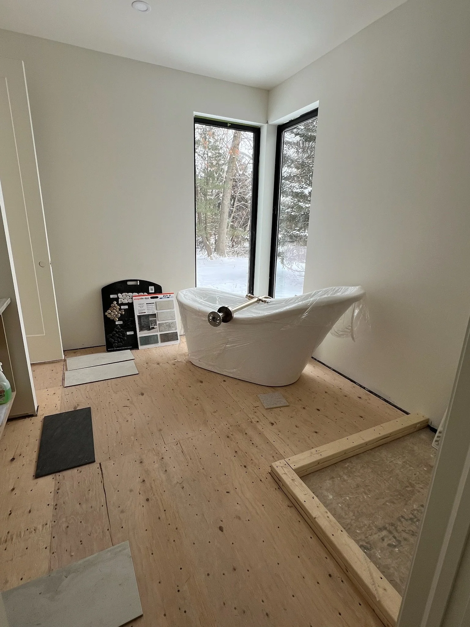 A bathroom under renovation with a freestanding bathtub covered in plastic, large windows, and construction materials on a wooden floor, with a snow-covered outdoor landscape visible through the windows.