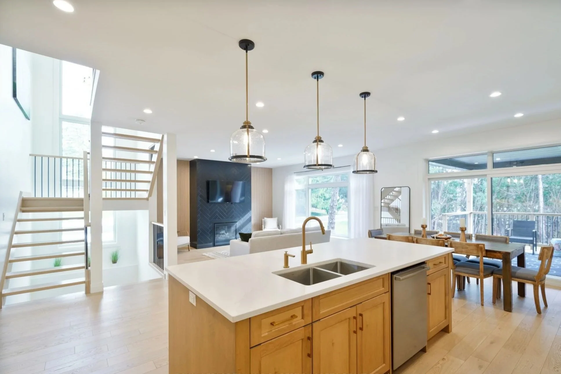 Open-concept living room and kitchen with island and dining area, large windows, and staircase leading upstairs.