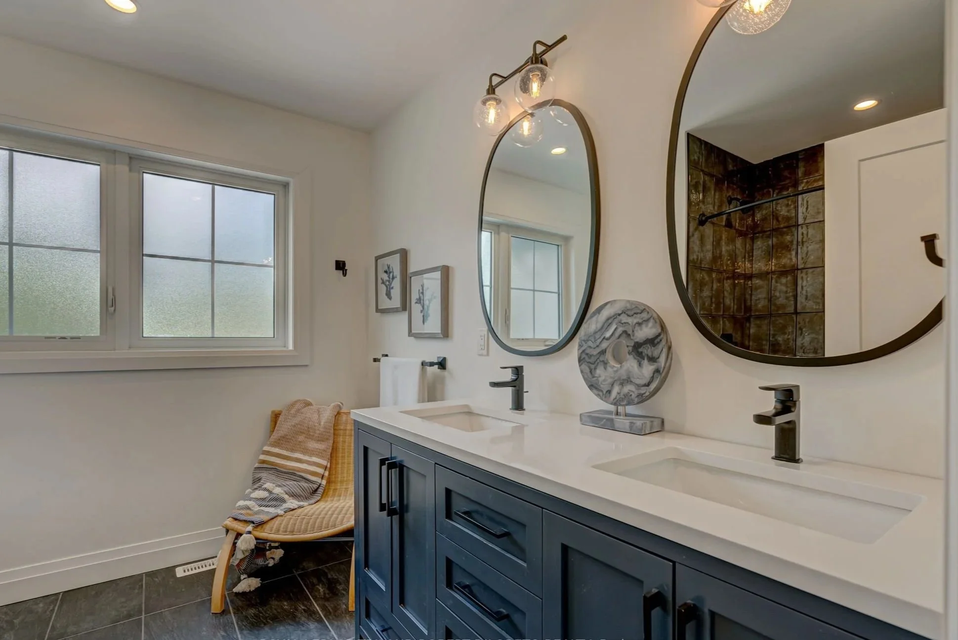 Modern bathroom renovation with dual sinks, navy blue vanity, two oval mirrors, a marble decorative object, two framed art pieces, frosted window, and a shower with brown tile walls.