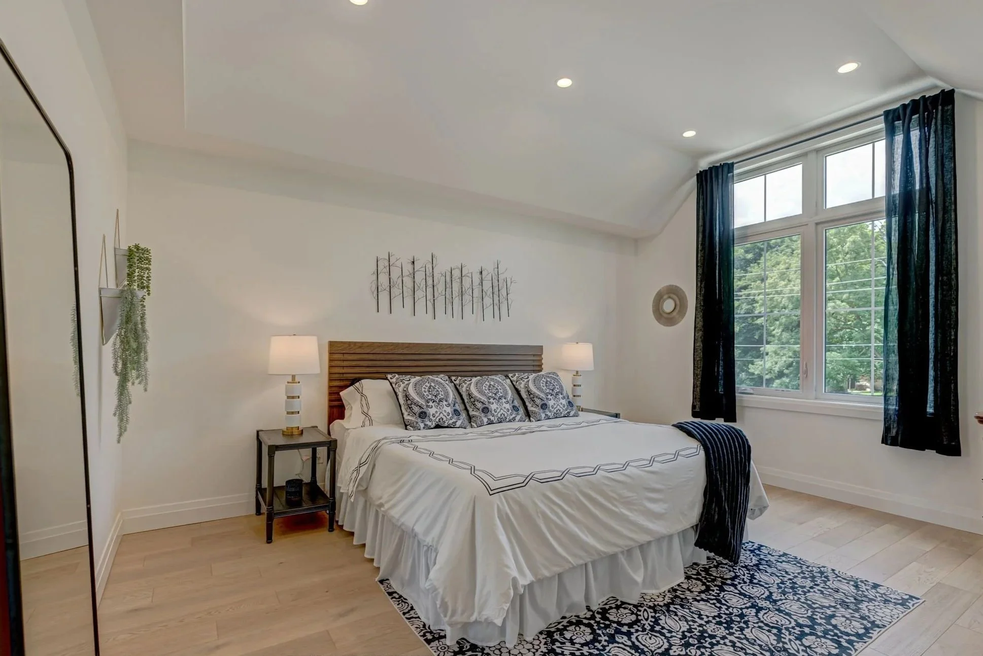 A bedroom with a large bed, white bedding, and decorative pillows, illuminated by natural light from a large window with black curtains, with side lamps on nightstands, a mirror, wall art, a black and white patterned rug, and green plants.