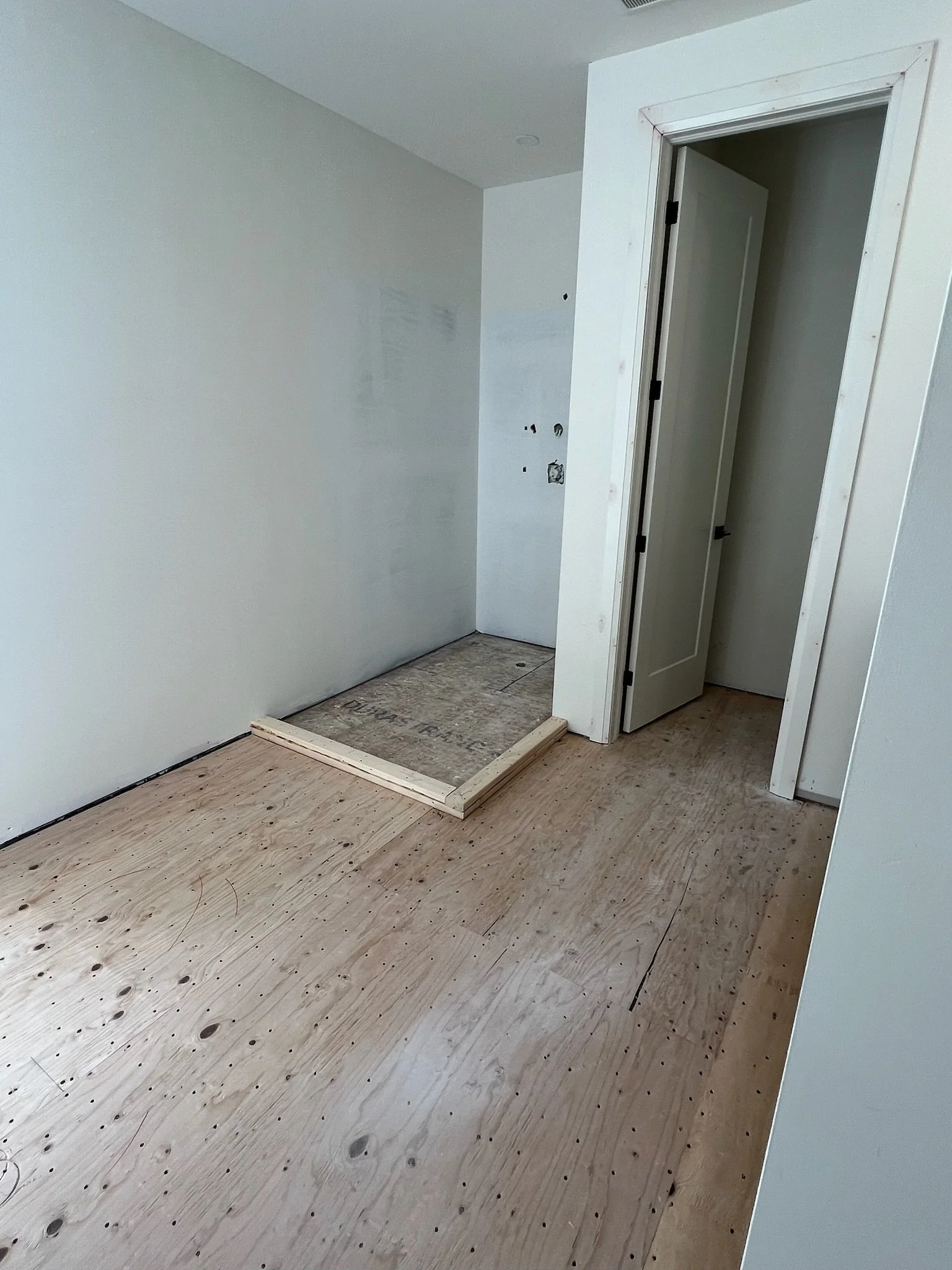 Bathroom renovation under construction with unfinished wooden flooring, an open doorway, and a small area with a raised platform.
