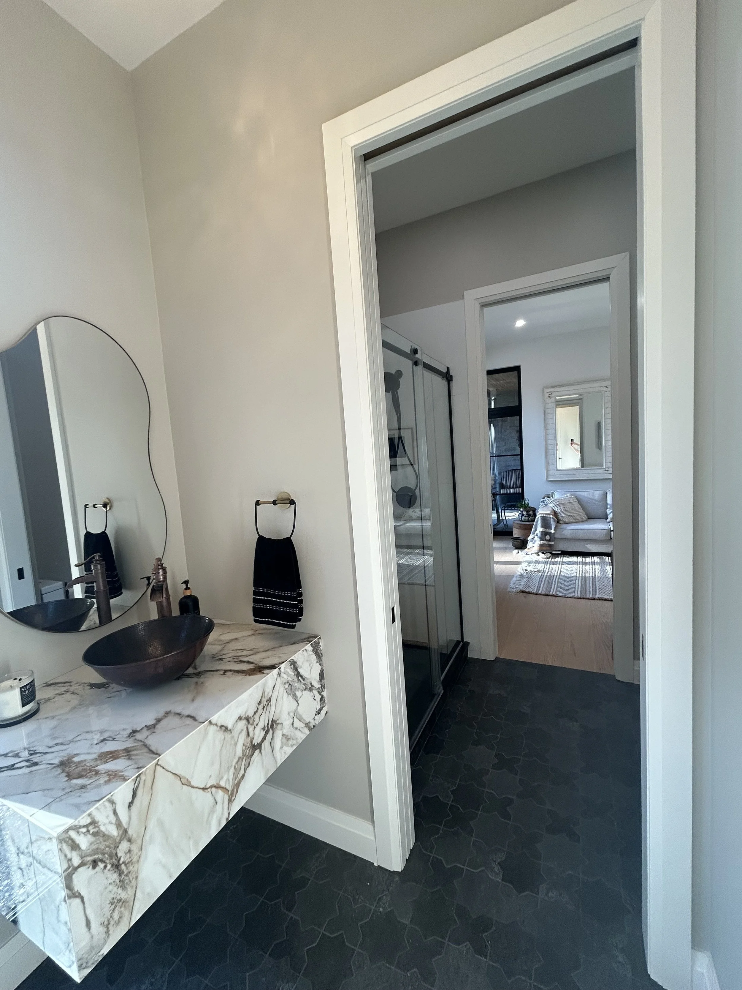 A bathroom renovation with a marble vanity, vessel sink, and round mirror. There are black towels on a towel ring, and a walk-in shower with a glass door. The bathroom opens into a living room with a sofa and window.