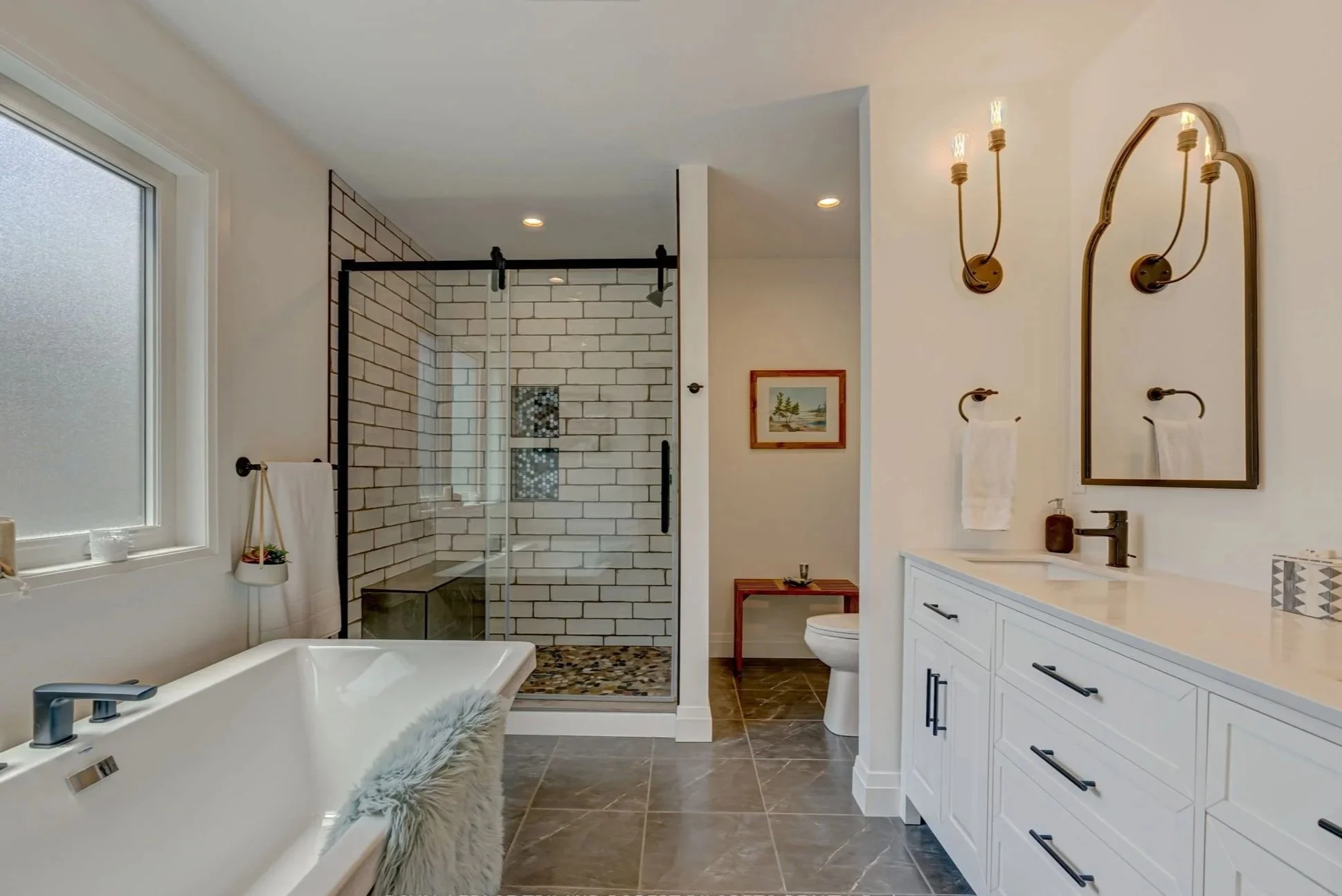 Modern bathroom renovation with a bathtub, a glass-enclosed shower with white brick tiles, a white vanity with black handles, and brass wall sconces above a mirror.
