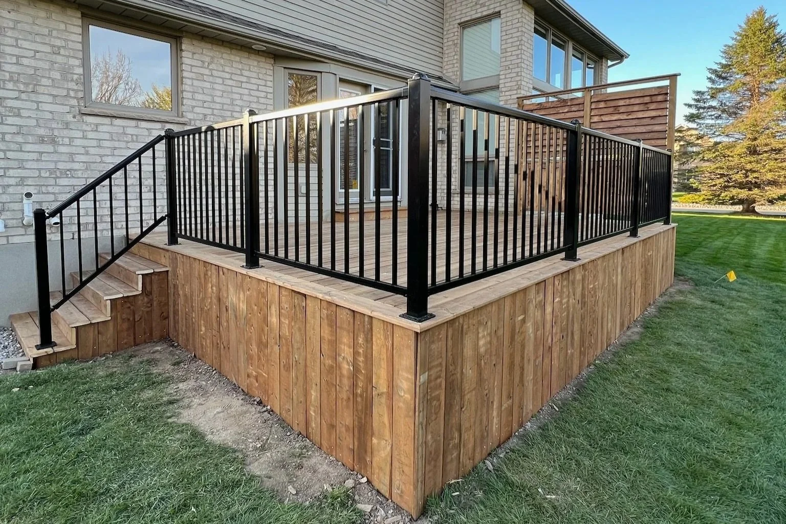 A wooden deck with black metal railing around it, attached to a house with brick walls and large windows, green lawn surrounding it, and a large tree in the background.