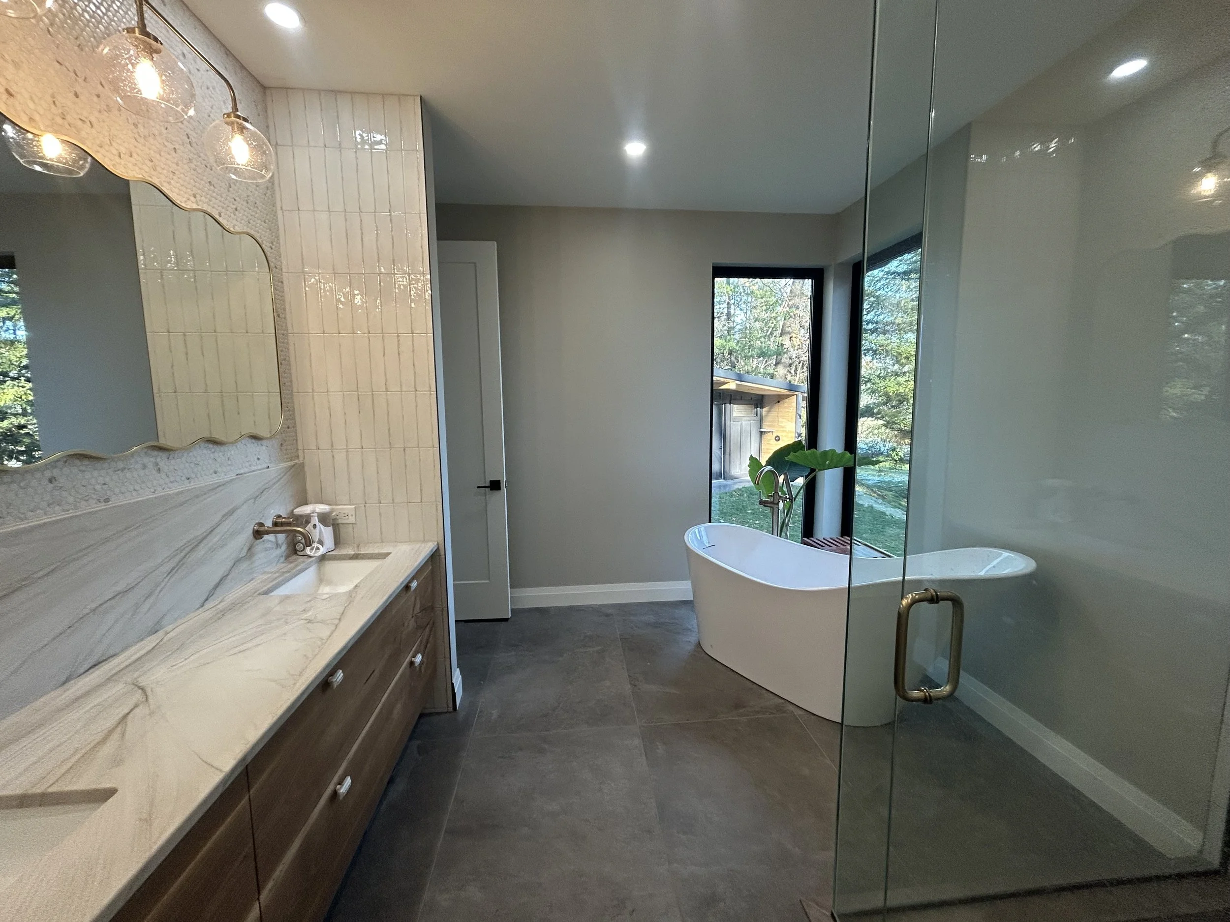 Modern bathroom featuring a white freestanding bathtub near a large window with trees outside, a wooden vanity with a marble countertop and sink, a decorative mirror, and a glass shower enclosure with a metal handle.