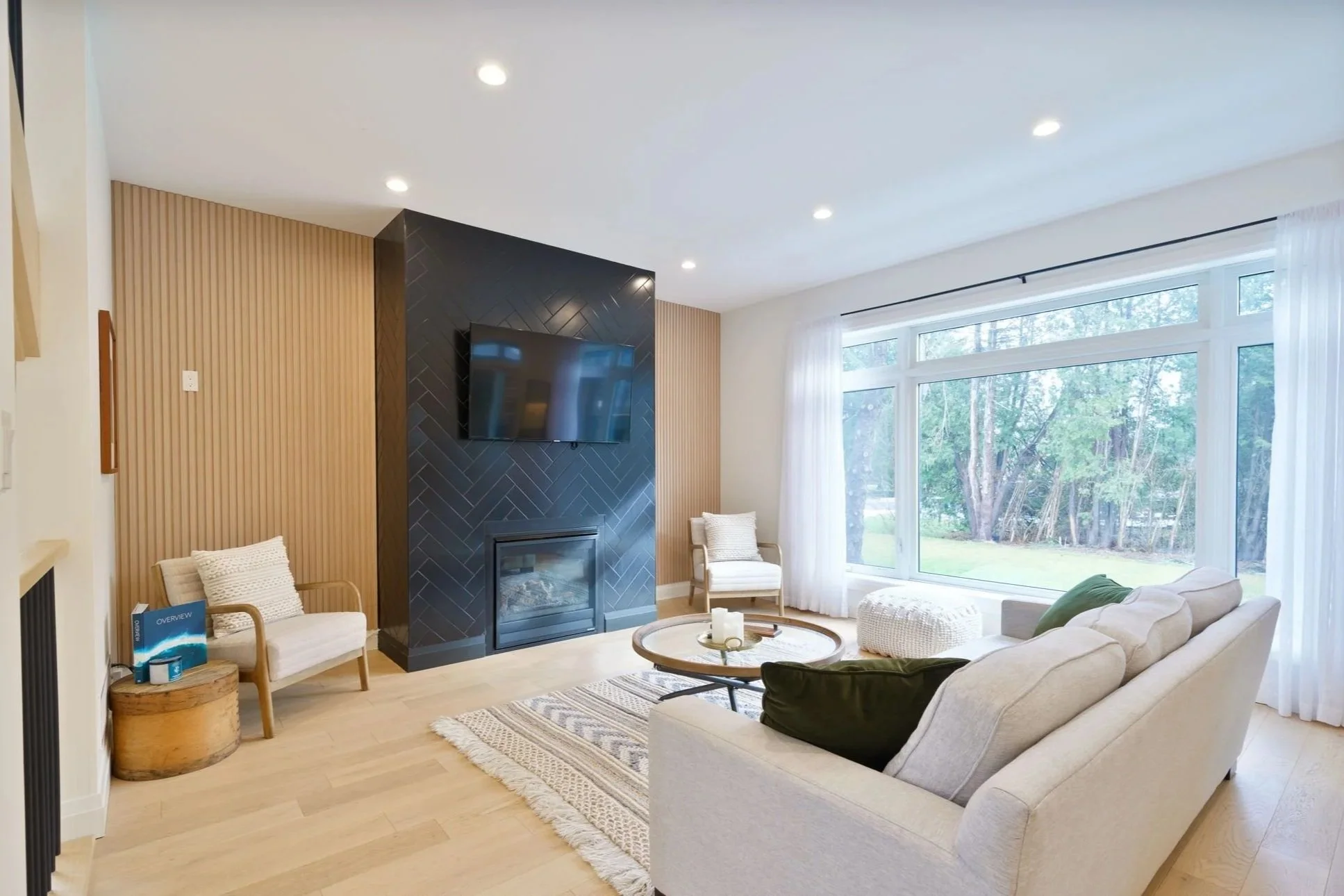 Bright living room with large window, beige sofa, armchairs, a black fireplace, and a wall-mounted TV.