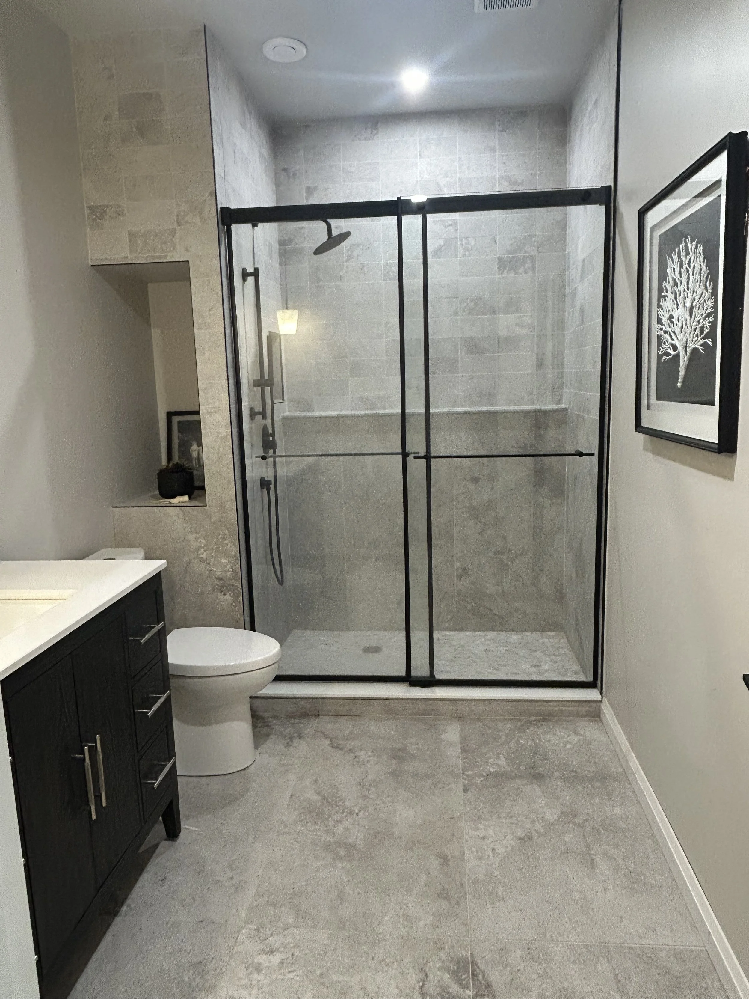 Finished bathroom renovation with a glass-enclosed shower, a black vanity with a white countertop, a toilet, a framed black-and-white art piece of a tree on the wall, and neutral-colored tiled flooring.