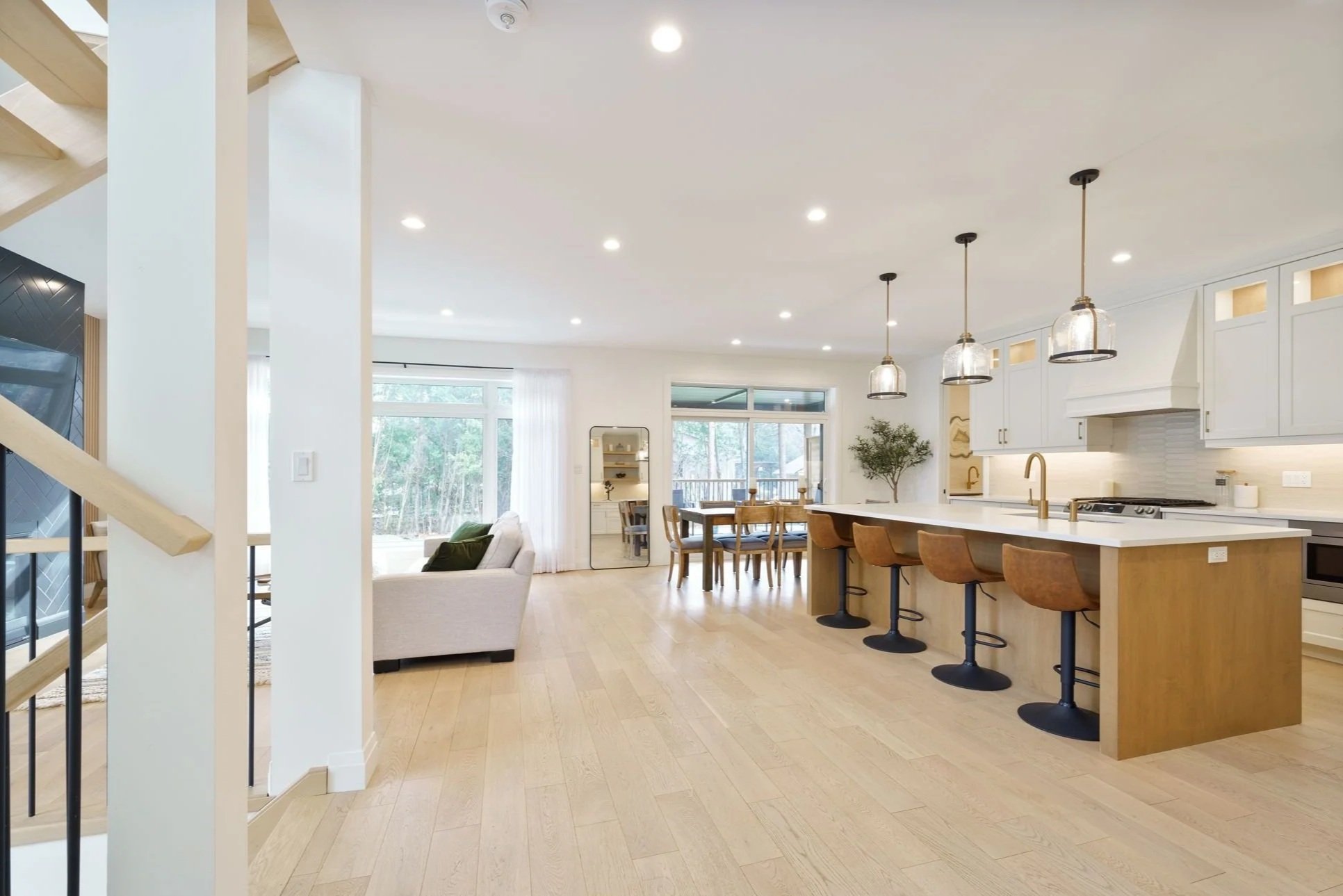 Open-concept kitchen and dining area with light wood floors, white walls, and modern furnishings, including a kitchen island with bar stools, dining table, and living room with sofa.