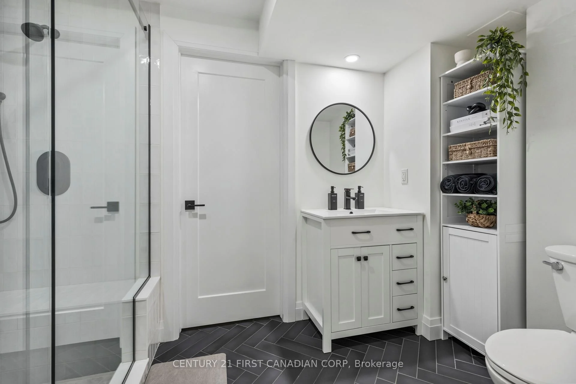 Modern white bathroom with black accents, including a black framed mirror, black cabinet handles, black towels, and black shower fixtures, with a glass shower enclosure and a single vanity with a round mirror and decorative plants.