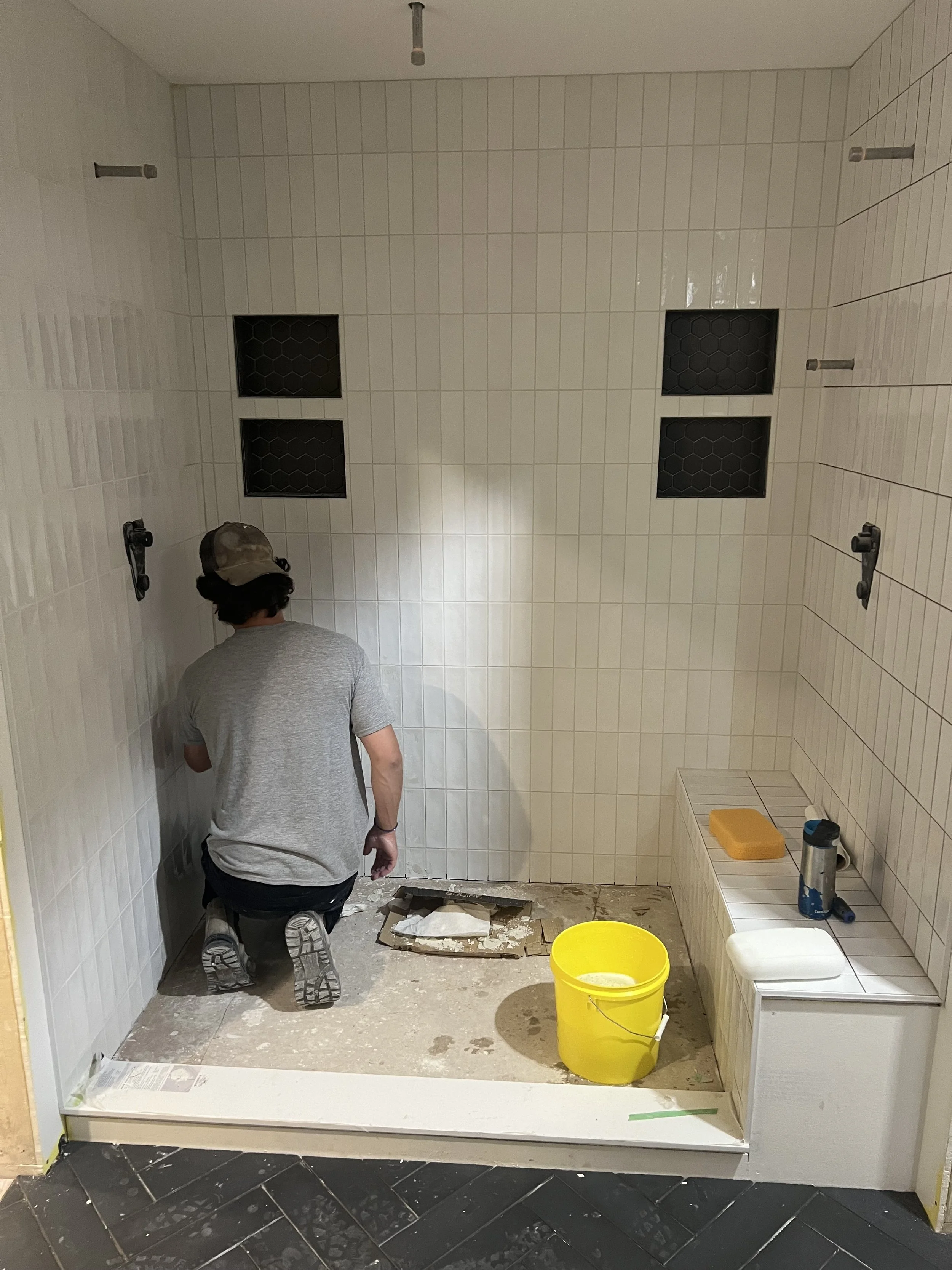 A person kneeling in a bathroom shower area under construction, with tiles on the walls, a yellow bucket, tools, and other construction materials present.