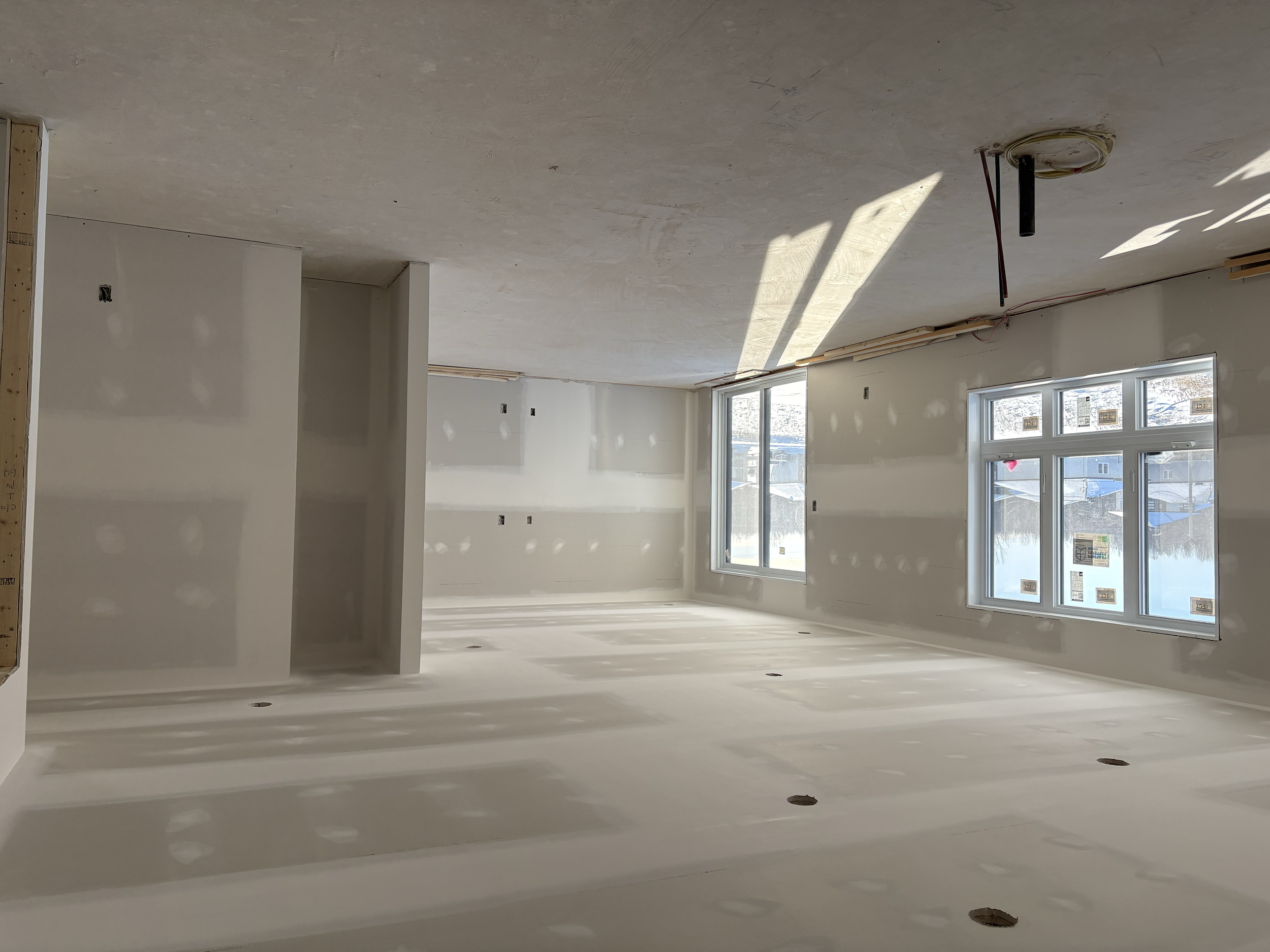 Intérieur d'un appartement encore en construction, avec des murs et un plafond en plâtre, deux fenêtres laissant passer la lumière et des trous au sol pour des prises ou un chauffage. Pas de meubles, en cours d'aménagement.