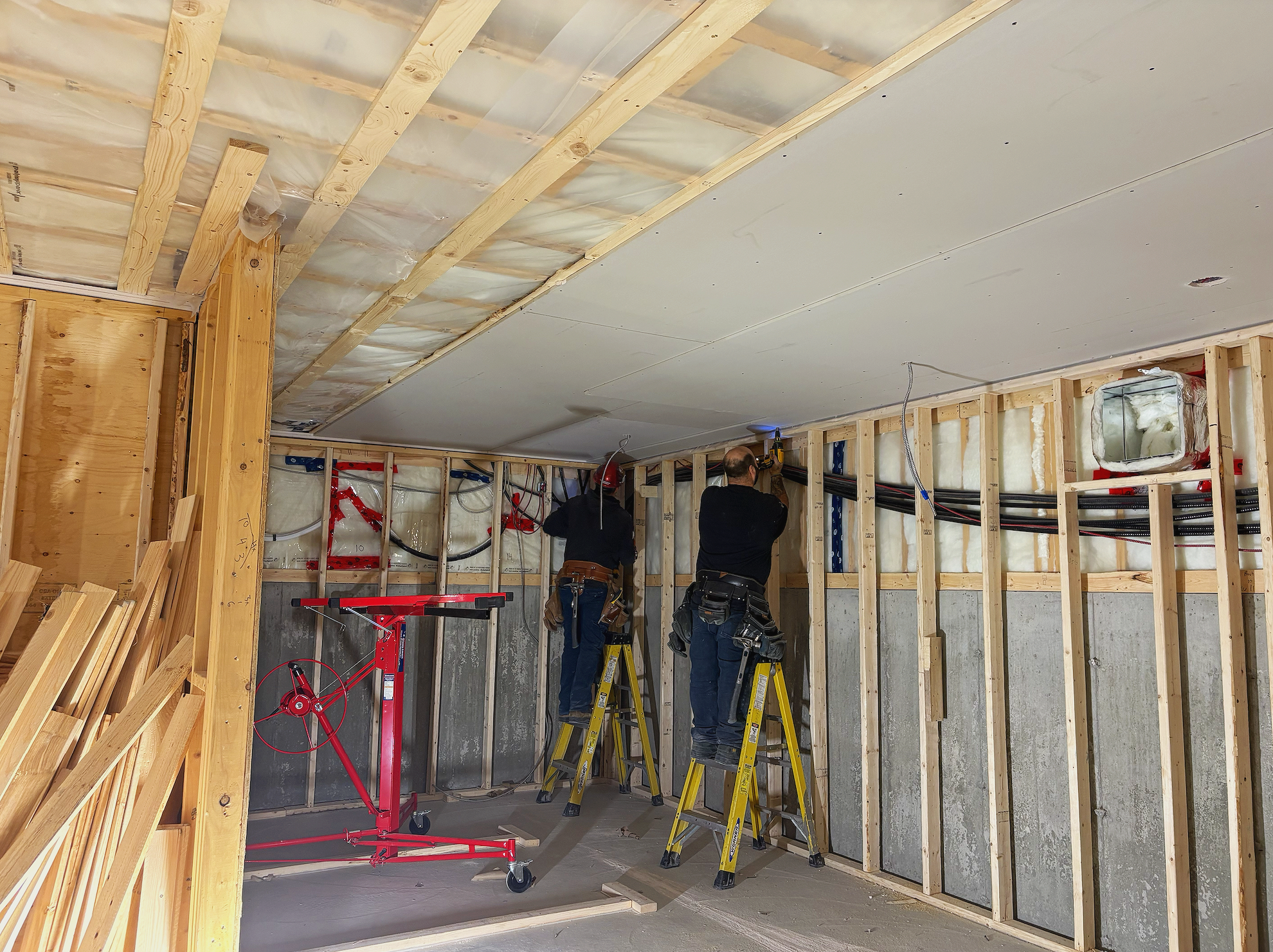 Deux ouvriers travaillent sur la construction intérieure d’un mur, utilisant des escabeaux et des outils, avec des murs en bois et des câbles électriques visible.